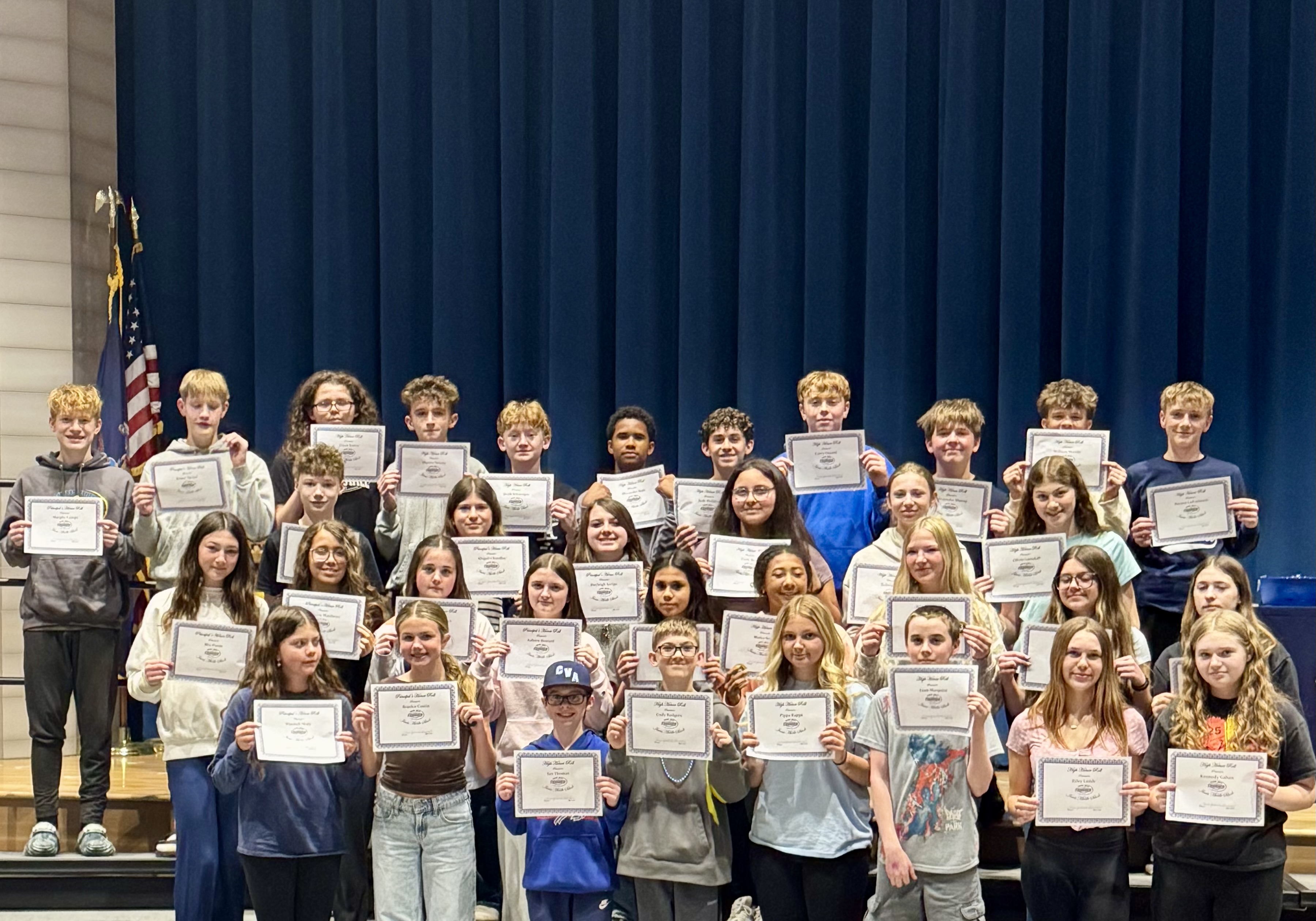A group of students proudly hold up certificates on a stage with a blue curtain backdrop.