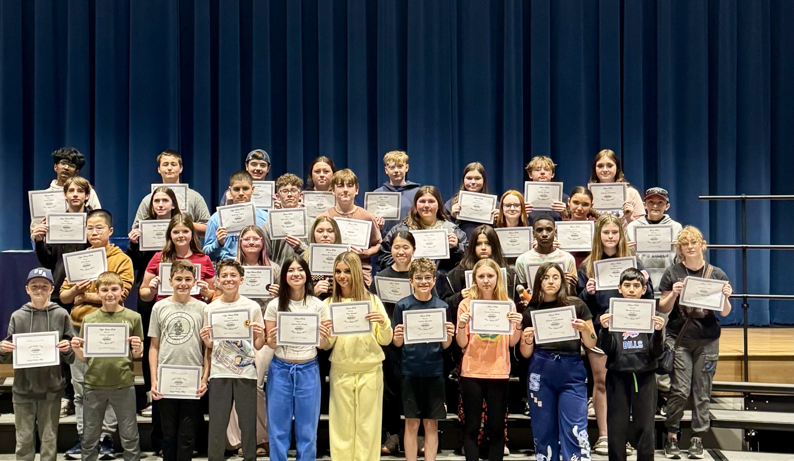 A diverse group of students stands on risers, holding certificates of achievement in front of a blue curtain.