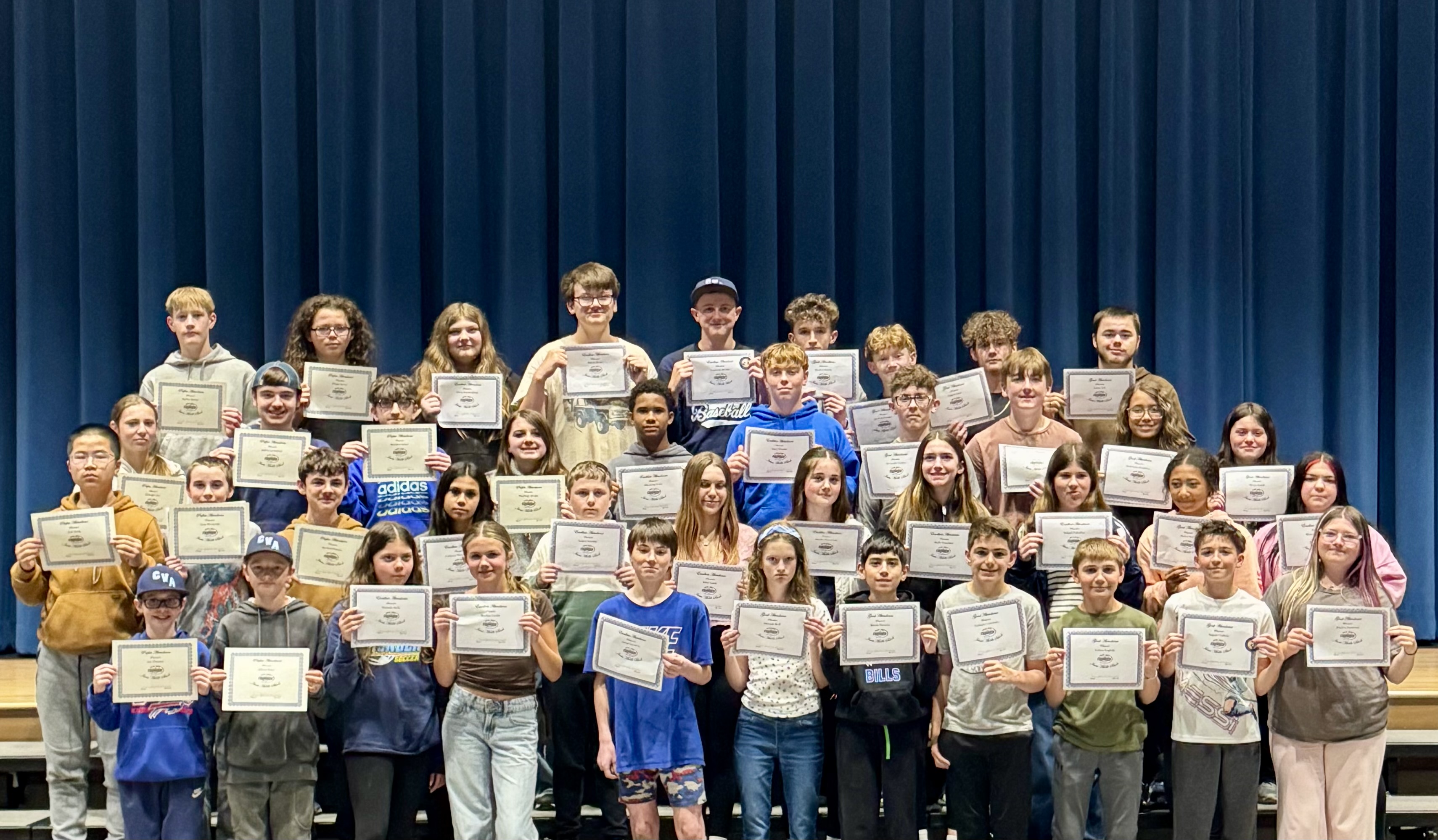 A large group of students stands on risers, holding certificates in front of a blue curtain.