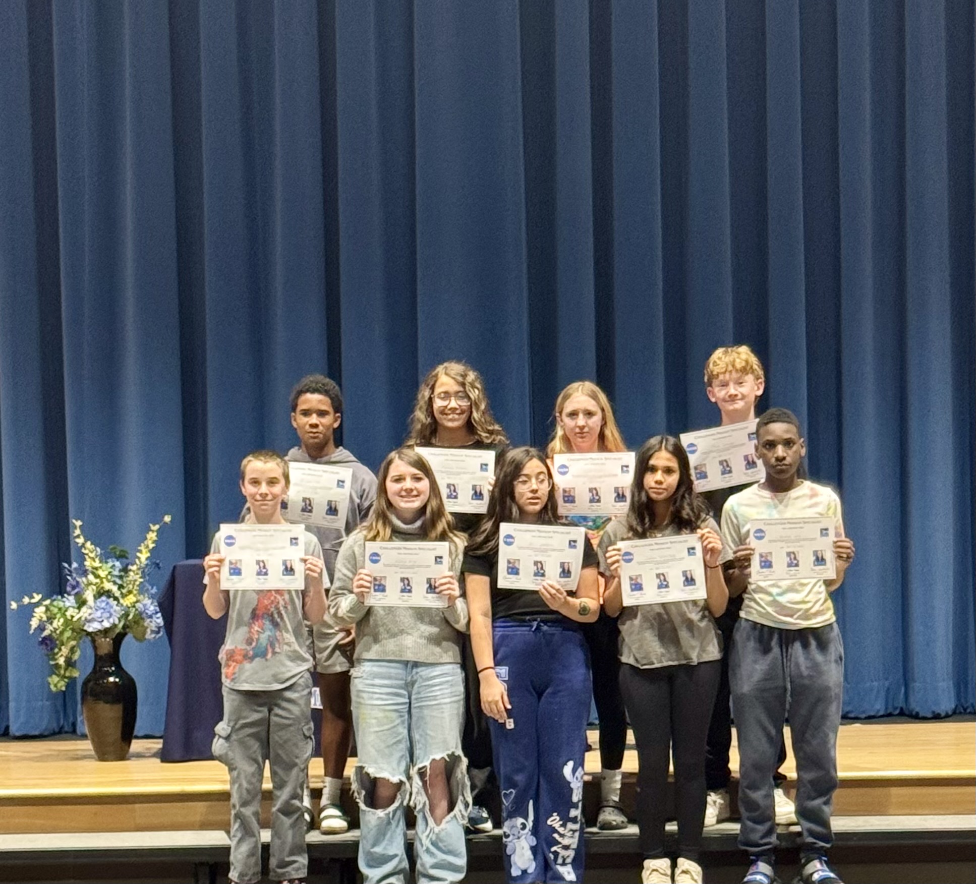 A group of nine students stand on a stage, holding certificates in front of a blue curtain.