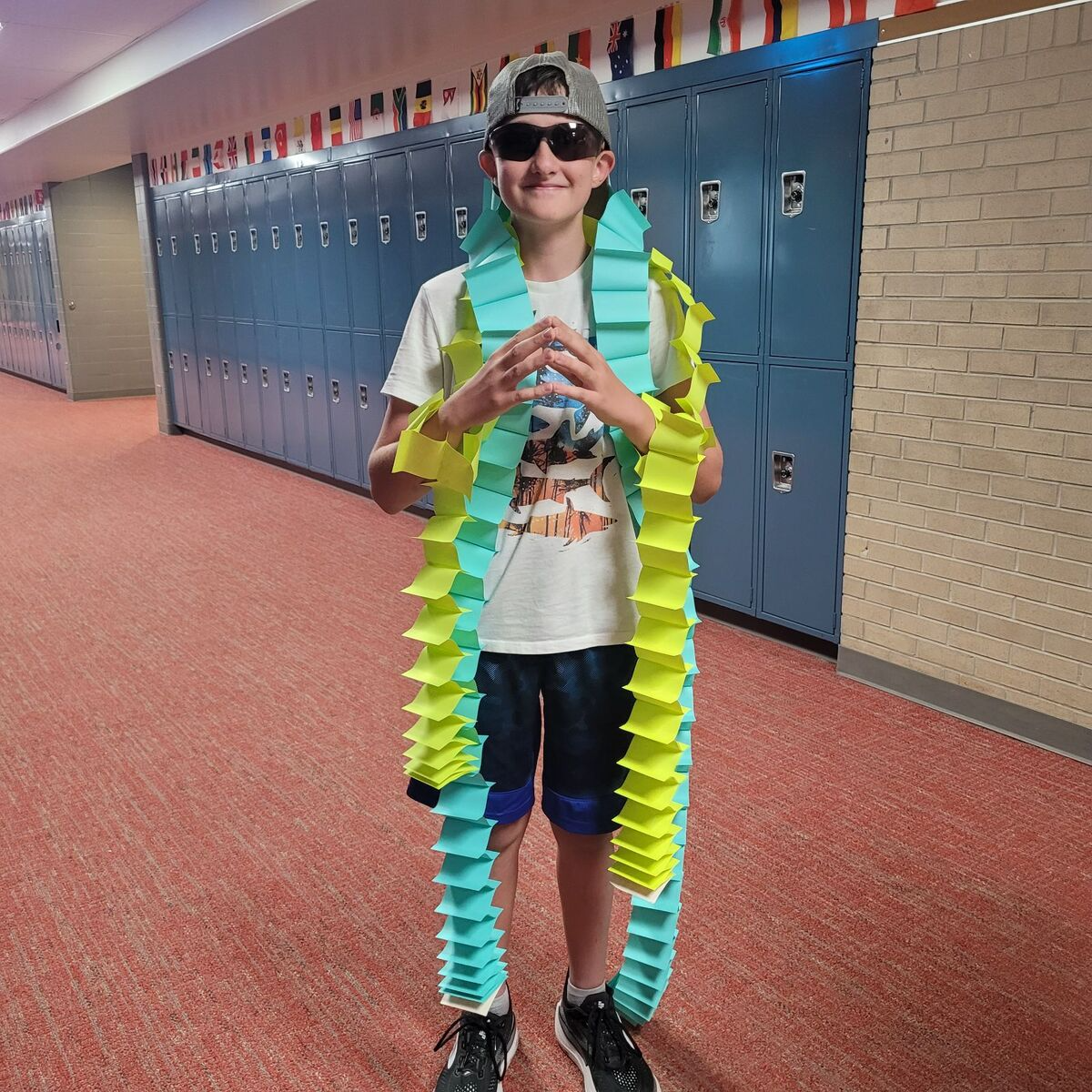 A young person stands in a school hallway, wearing a long, colorful garland made of folded paper.