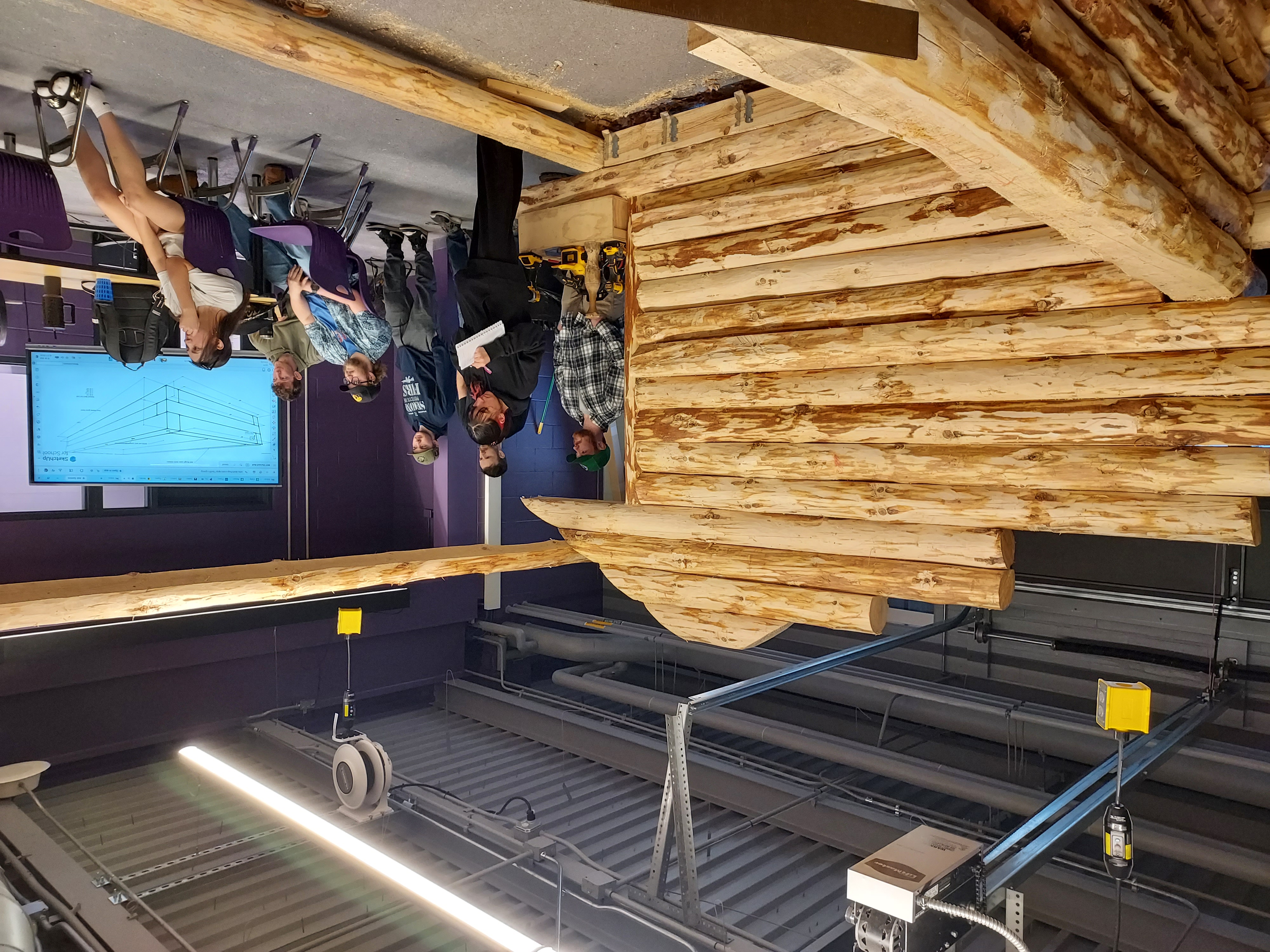 Students observe a log cabin structure being built in a workshop setting.