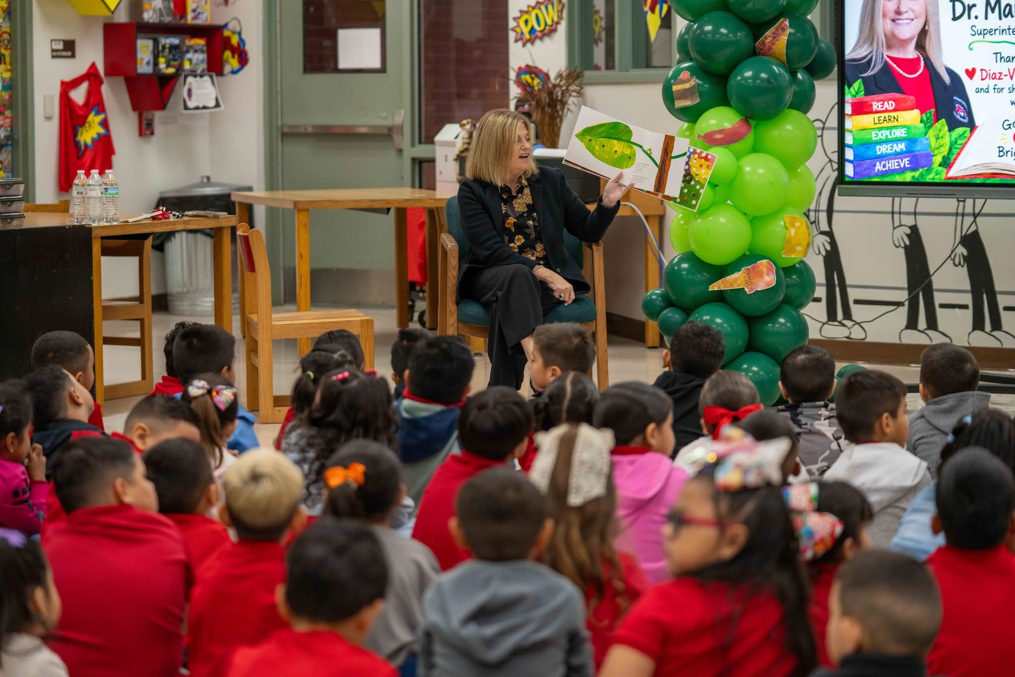 A woman reads a large picture book to a group of young children sitting on the floor in a classroom.