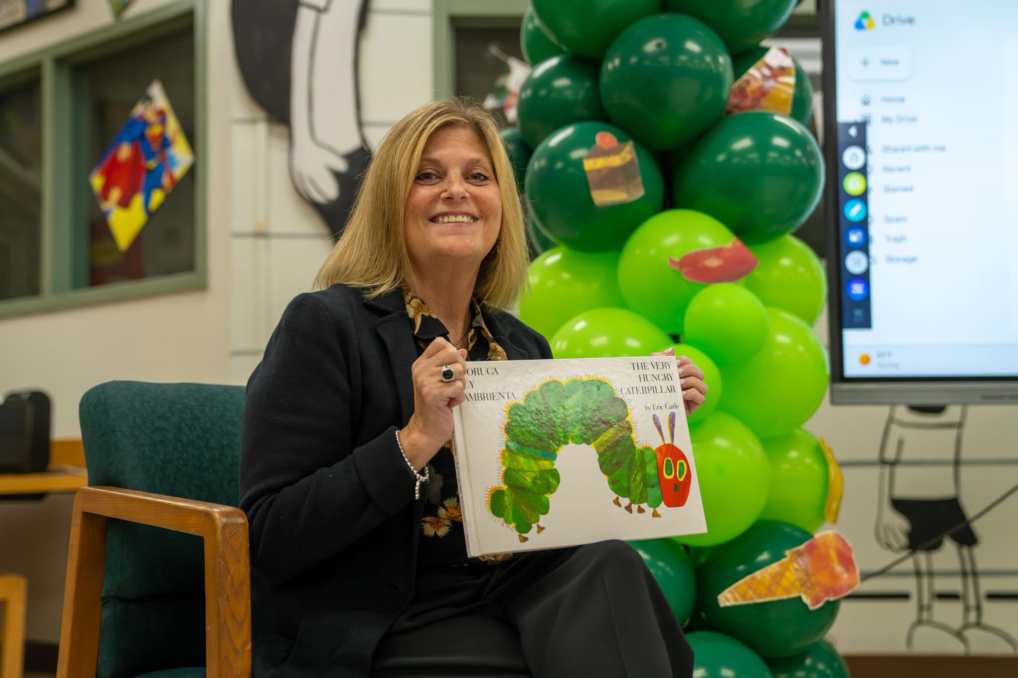 A smiling woman holds up "The Very Hungry Caripillar" book in a classroom with green balloon decorations.