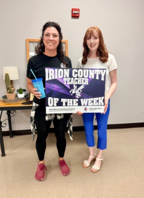 Two women stand together holding a sign that reads 'IRION COUNTY TEACHER OF THE WEEK'.