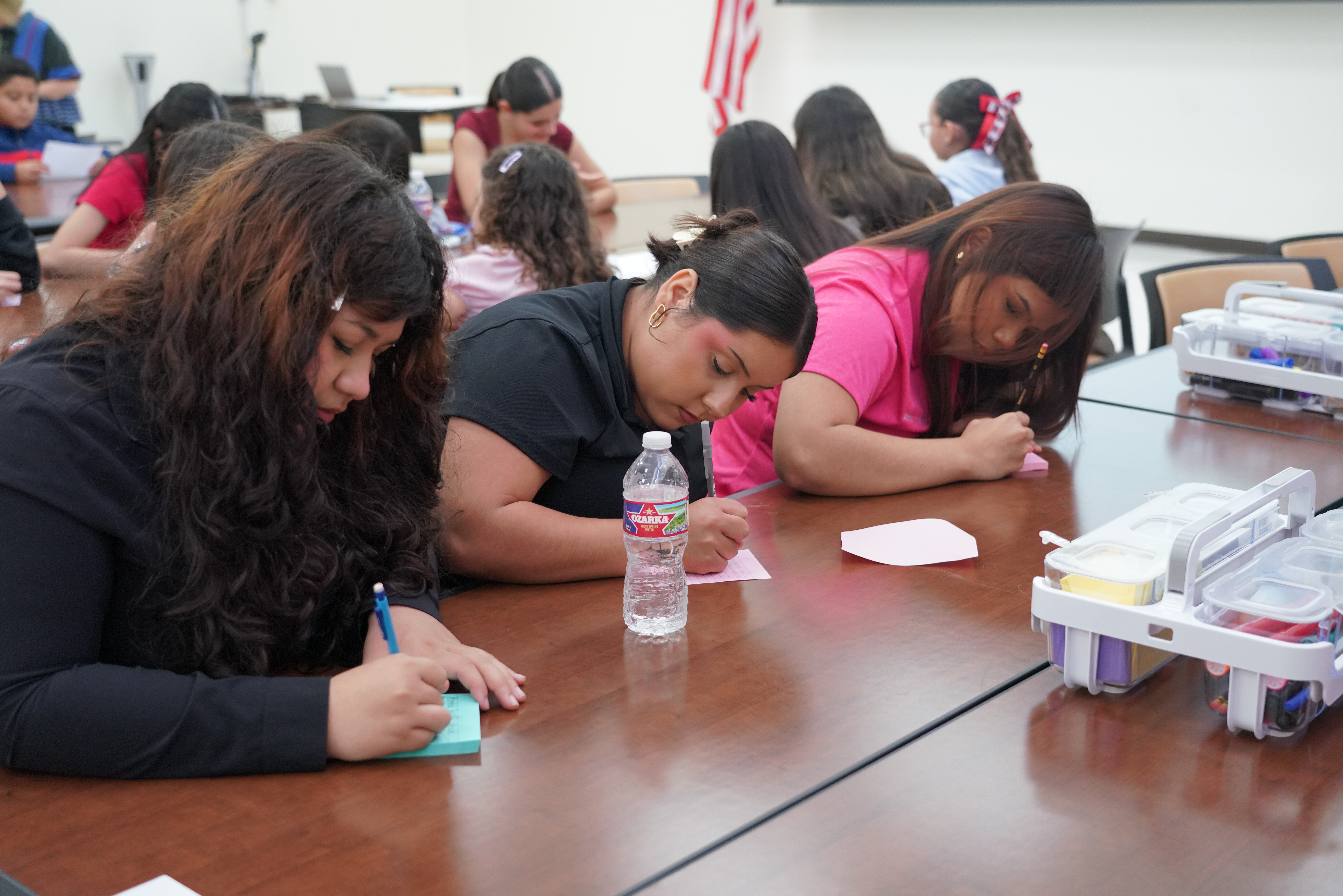 Three young women sit at a long table indoors, intently writing on small sticky notes.