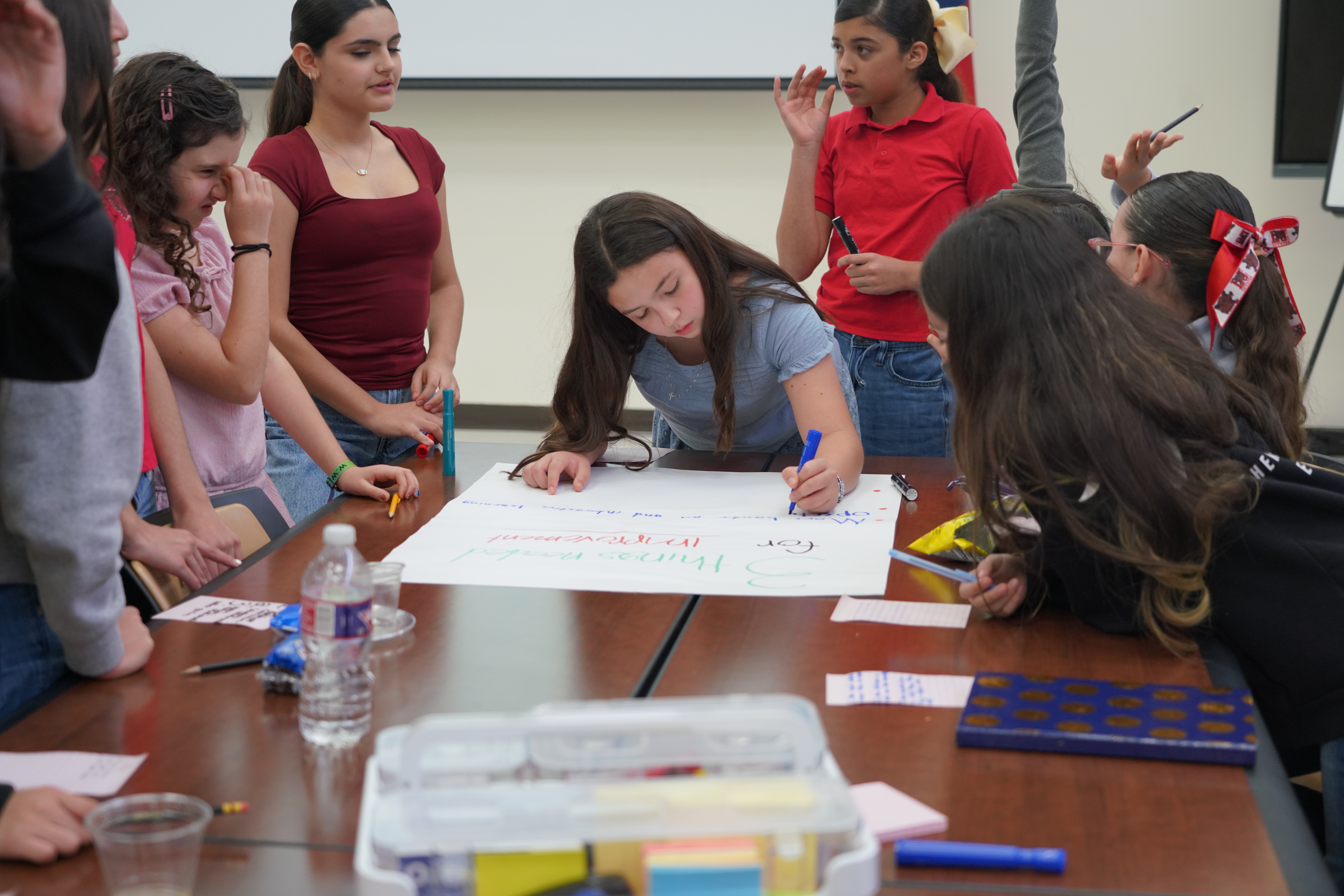 A group of students gather around a large table, collaborating on a project with markers and paper.