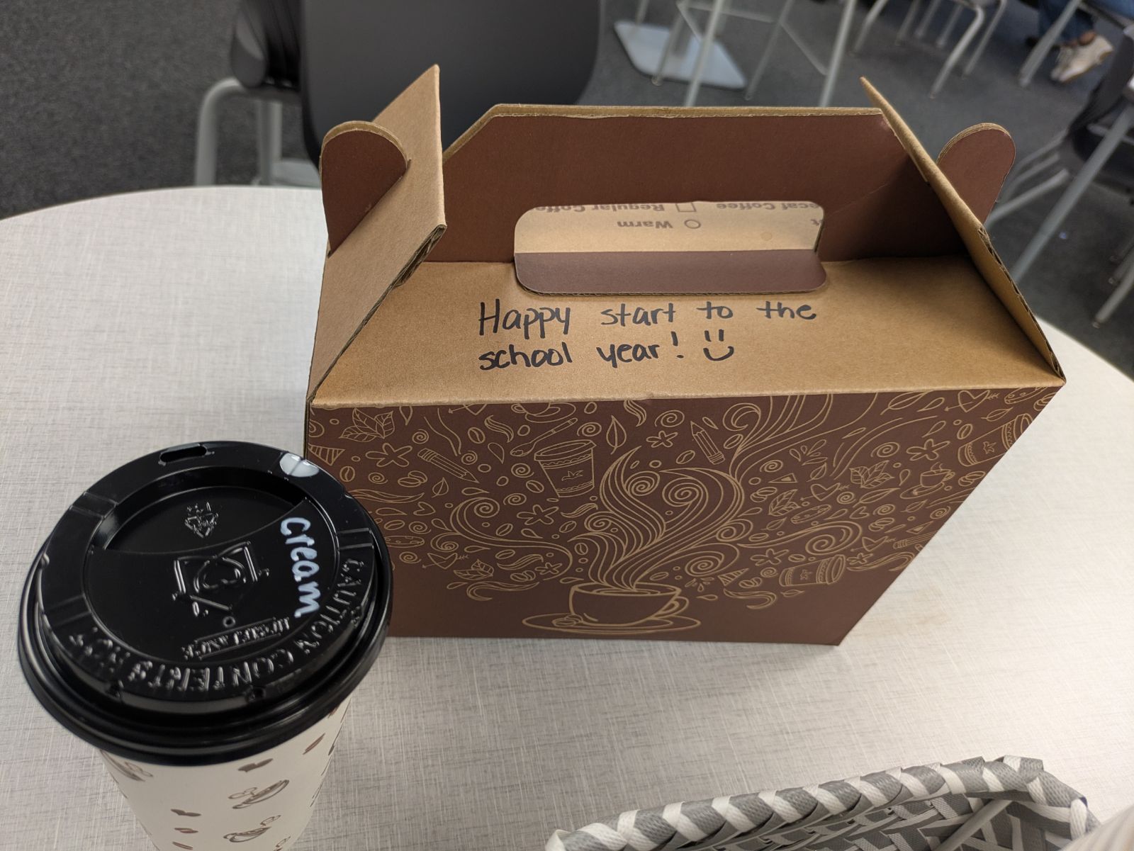 A brown cardboard box with a handwritten message 'Happy start to the school year!' sits next to a coffee cup.
