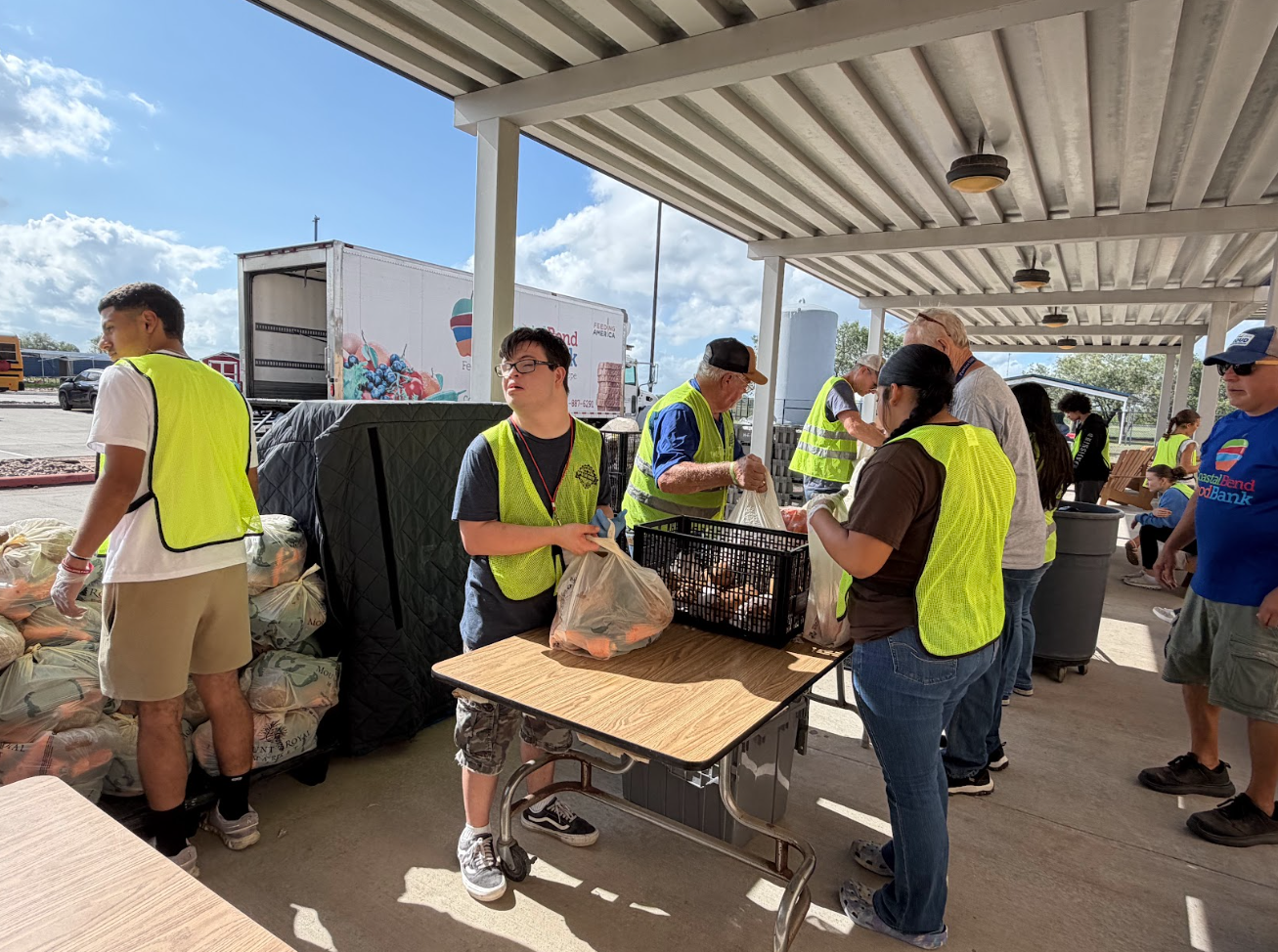 Volunteers in yellow safety vests pack bags of food at an outdoor community food bank distribution event.