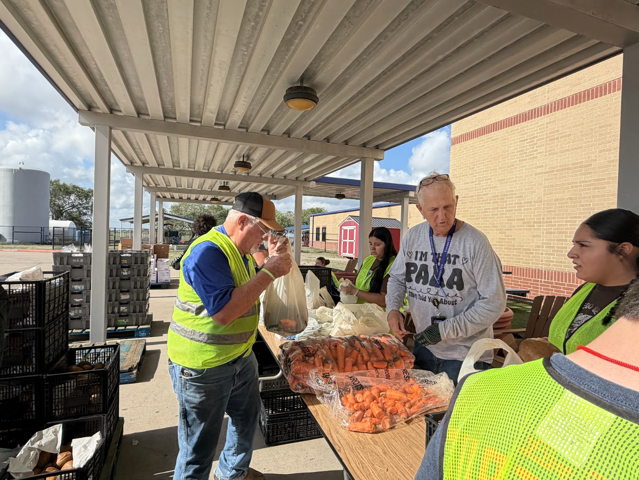Volunteers pack bags of fresh carrots outdoors under a covered walkway at a community food drive.