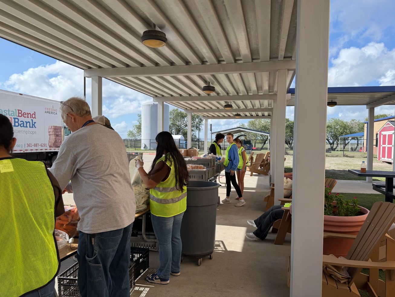 Volunteers wearing yellow safety vests sort and distribute food items outdoors under a covered pavilion near a food bank truck.