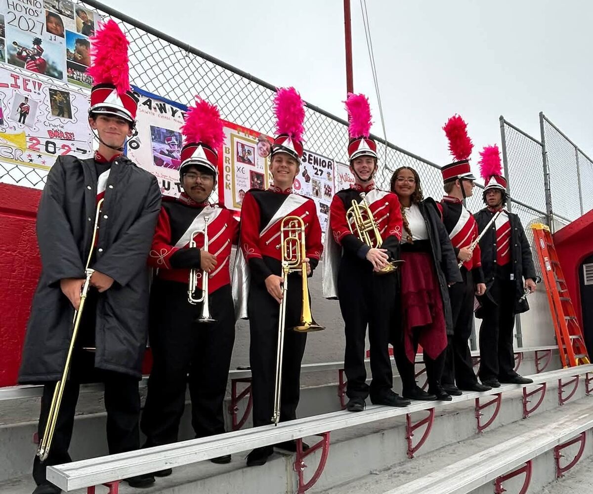 A marching band stands on bleachers in red, black, and white uniforms with tall pink plumes on their hats.