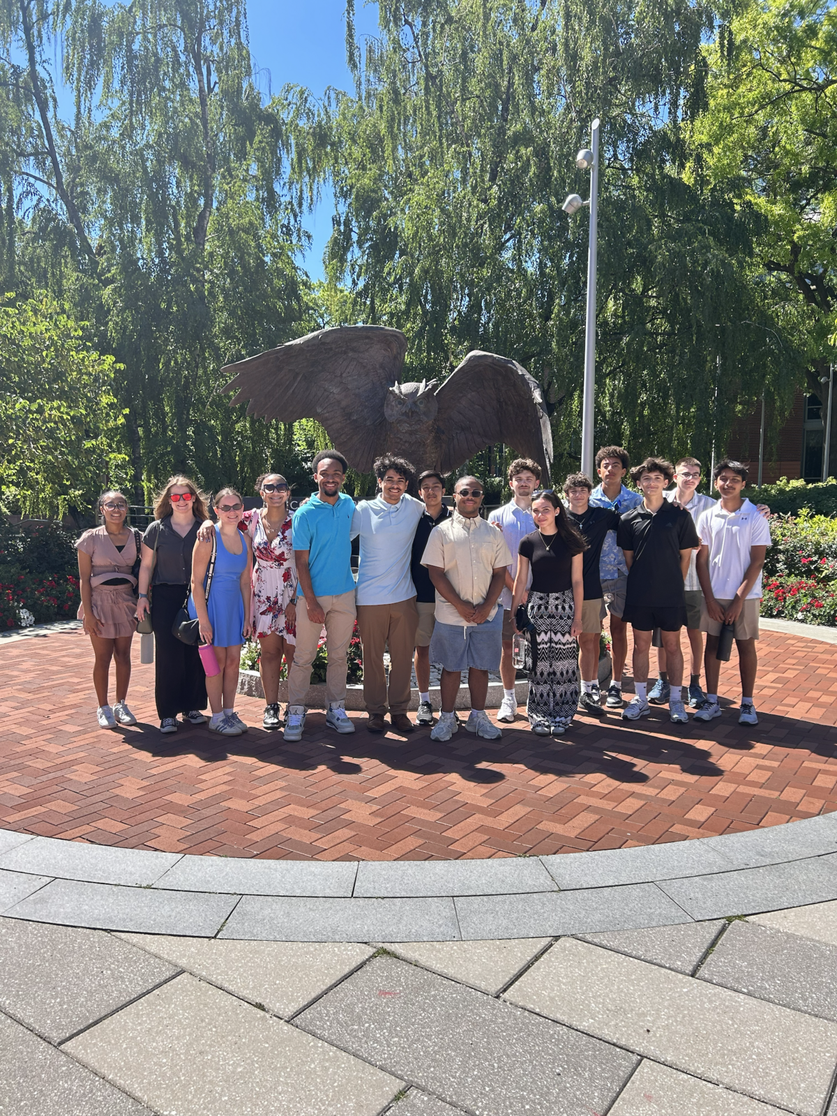 A diverse group of young people stand together outdoors in front of a large owl sculpture.