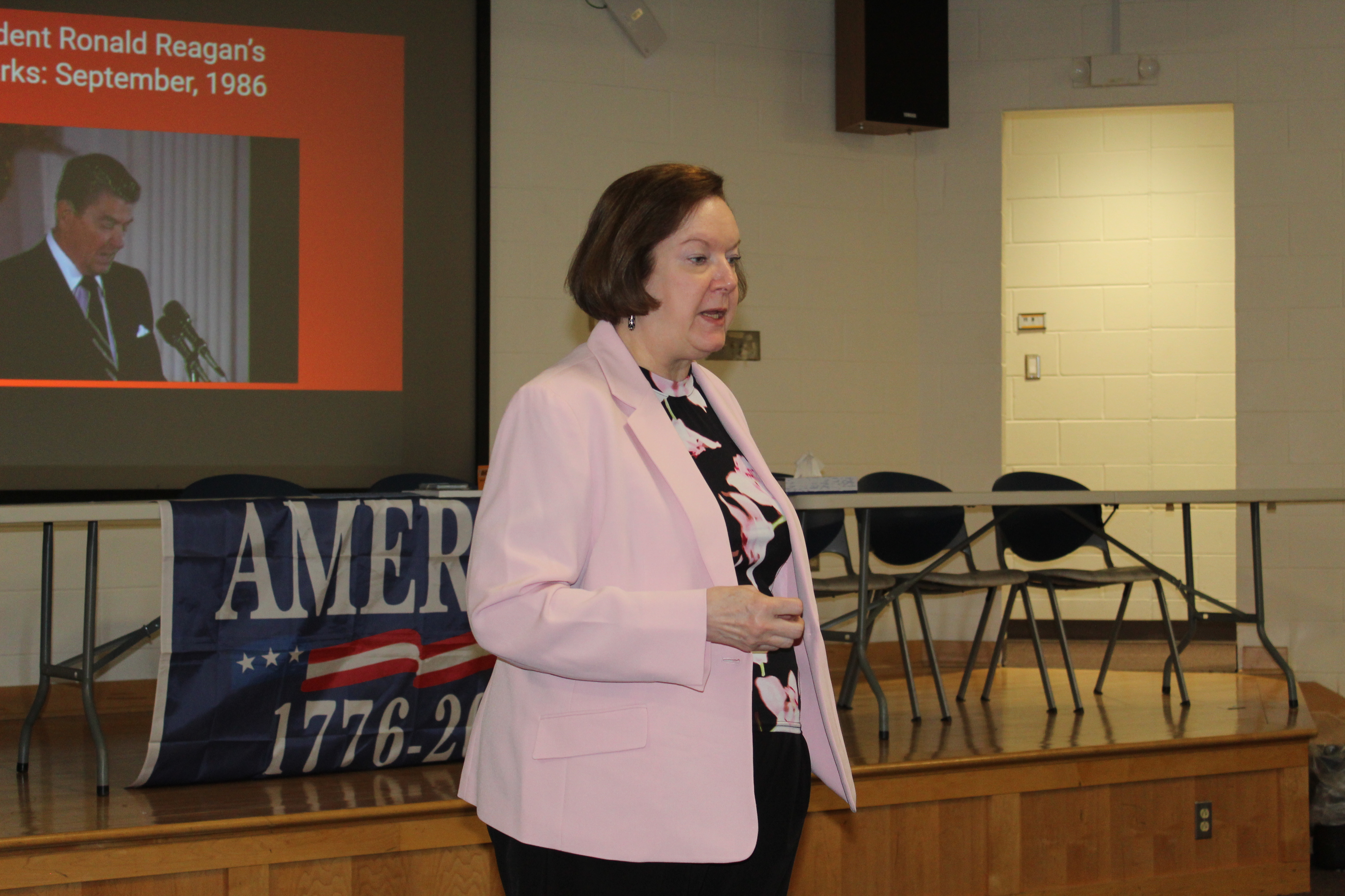 A woman in a pink blazer speaks at a podium in front of a projection screen showing President Ronald Reagan.