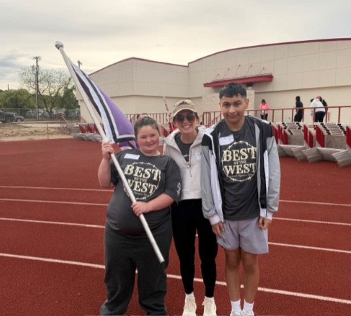 Three people stand on a red track, two wearing shirts that say "BEST OF THE WEST" and one holding a purple and white flag.