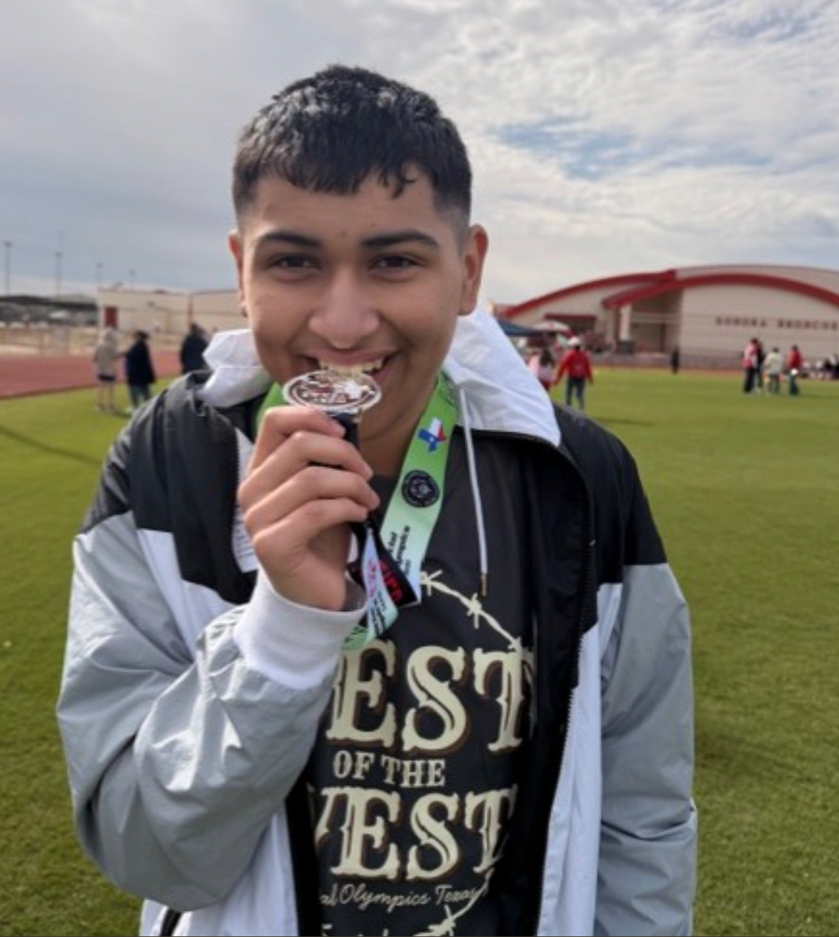 A young person smiles proudly, holding a medal near their mouth on a grassy field.