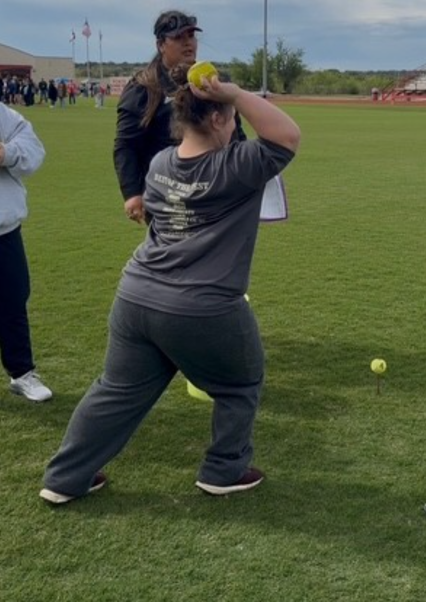 A person in a grey t-shirt and dark pants prepares to throw a yellow softball on a grassy field.