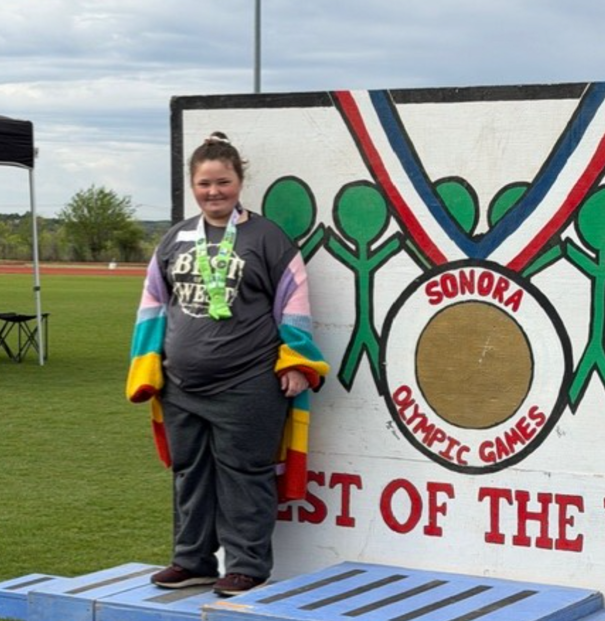 A young person stands on a blue podium in front of a mural depicting a medal and stick figures, wearing a medal.