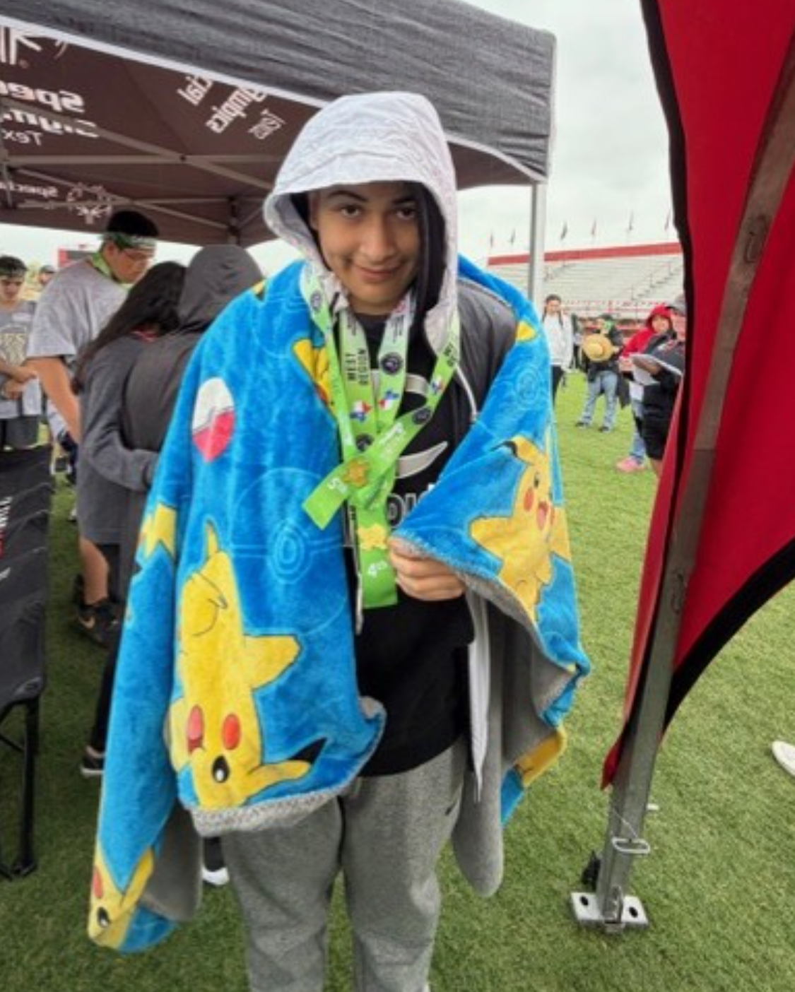 A young person wearing a Pikachu blanket and medals smiles at the camera.
