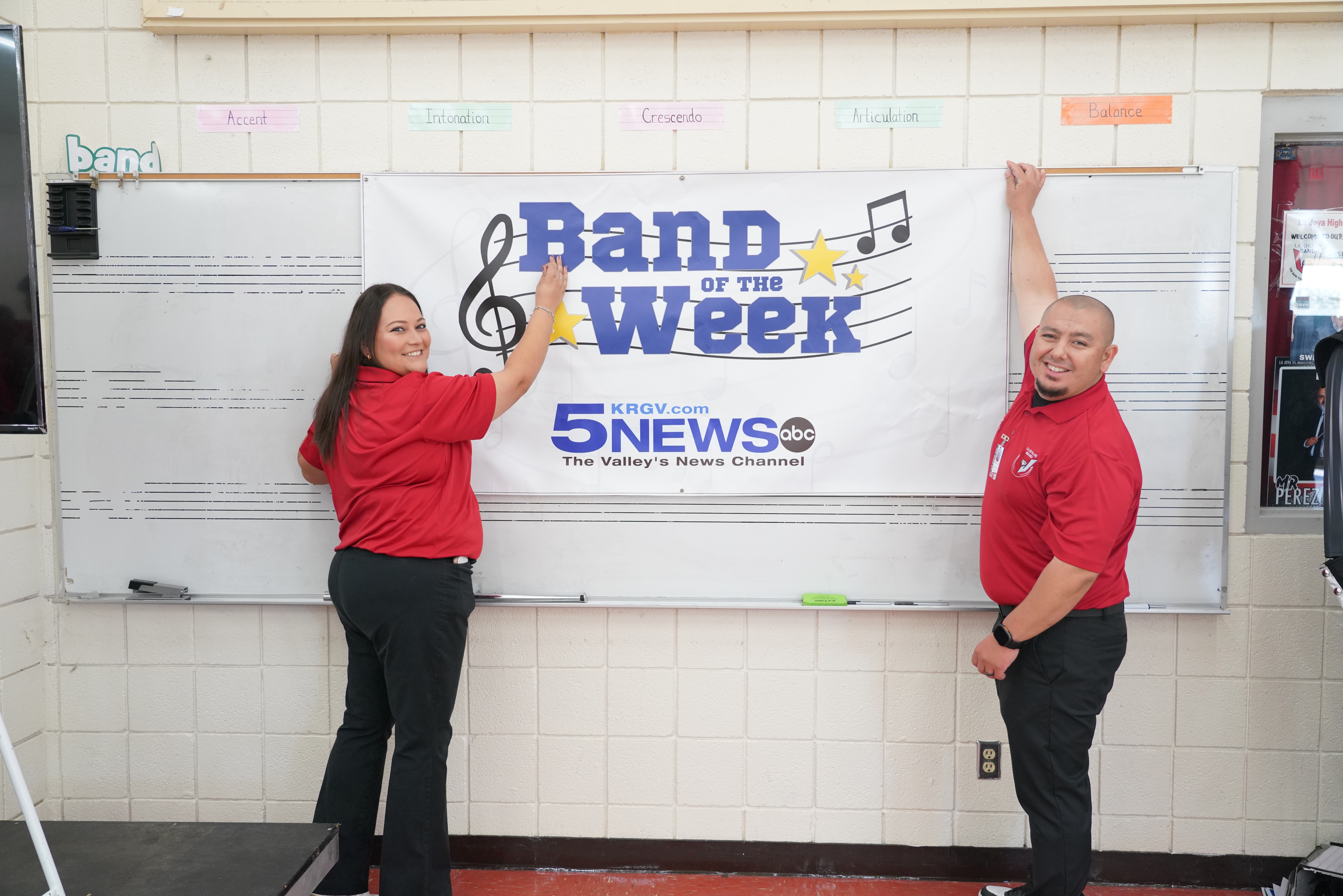 Two teachers in red shirts proudly hold up a banner that reads "Band of the Week" in a classroom.
