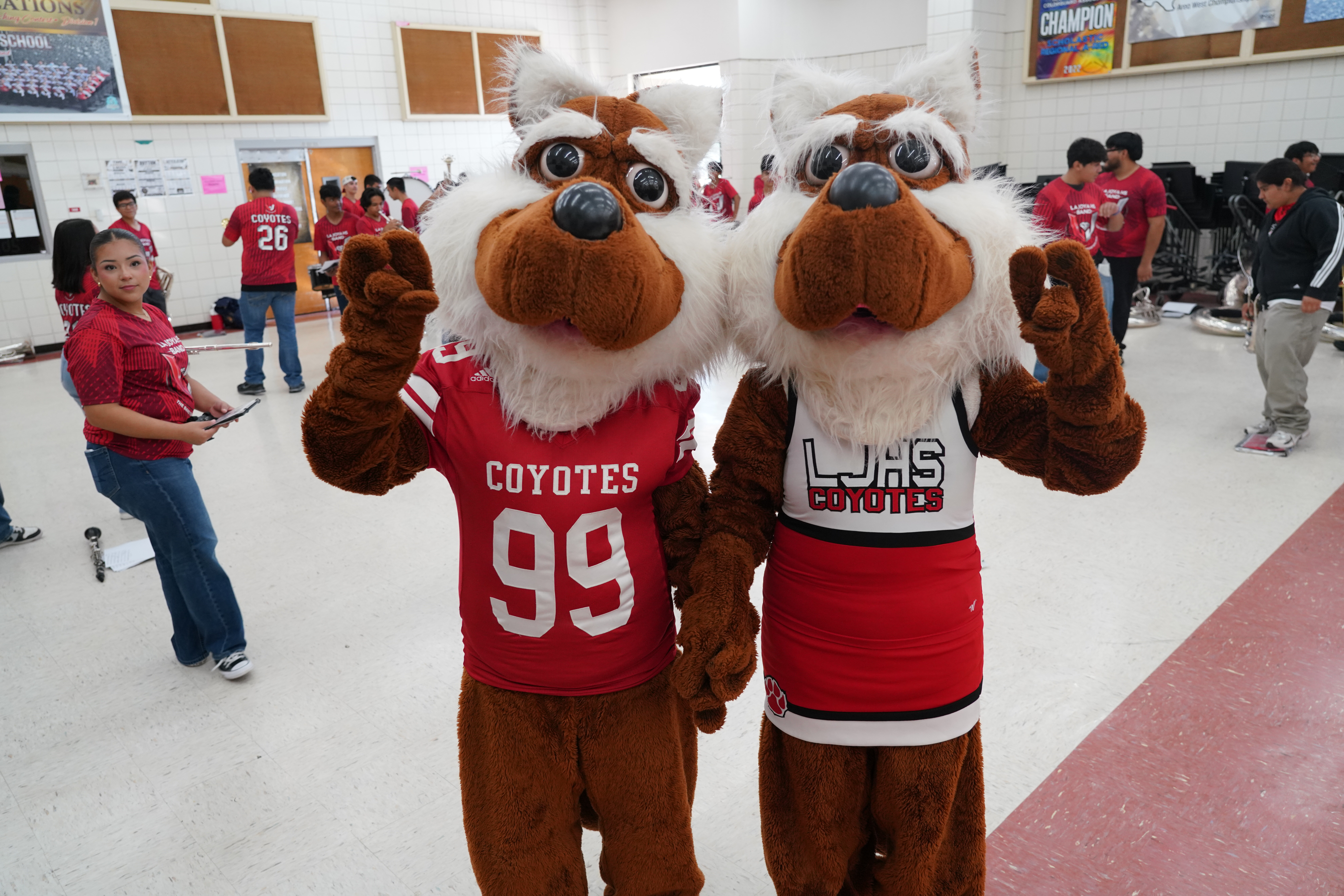 Two coyote mascots in red and white uniforms pose together in a school gym.
