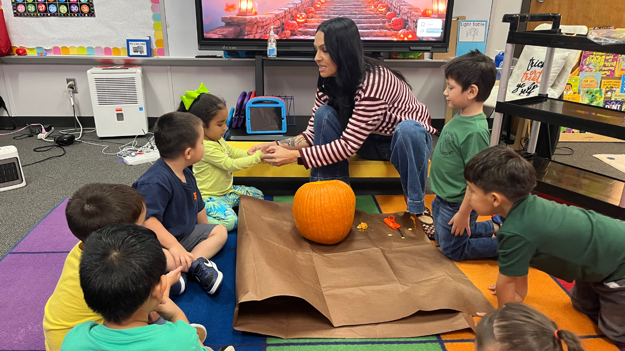 A teacher sits with a group of young students in a classroom gathered around a large orange pumpkin on the floor.