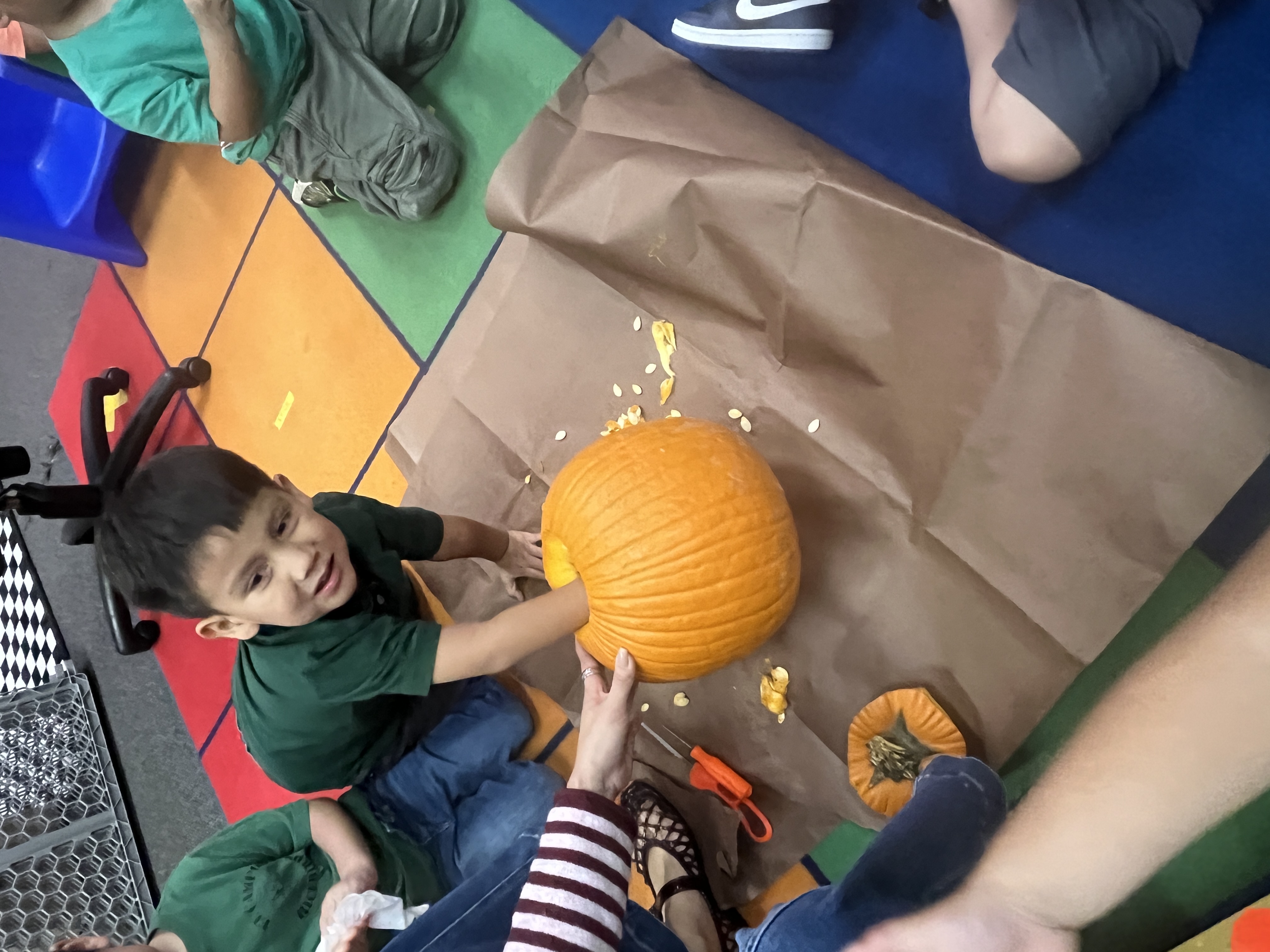 A smiling child reaches inside a hollowed-out pumpkin on a brown paper-covered floor.