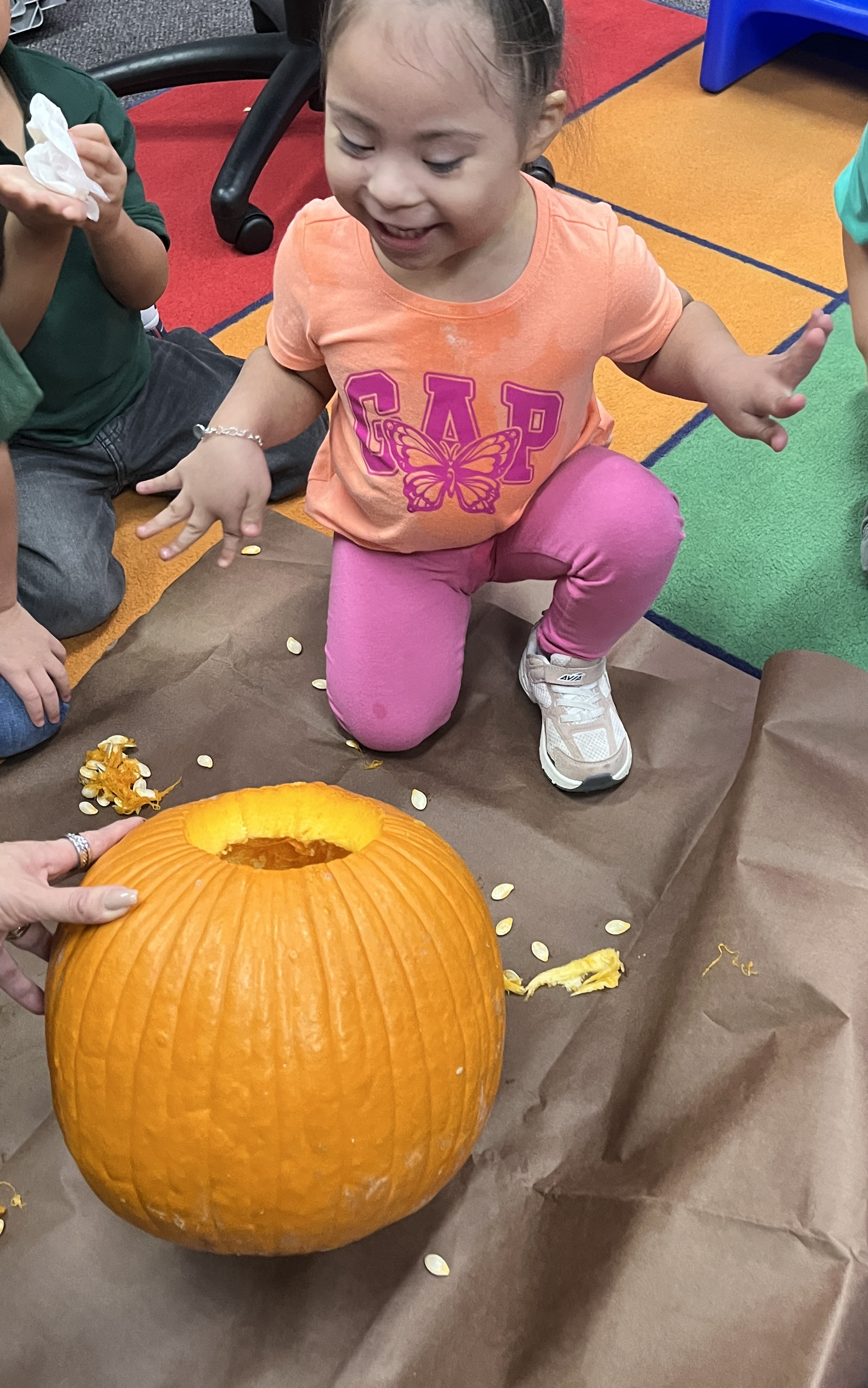A smiling child kneels next to a carved pumpkin on brown paper indoors.