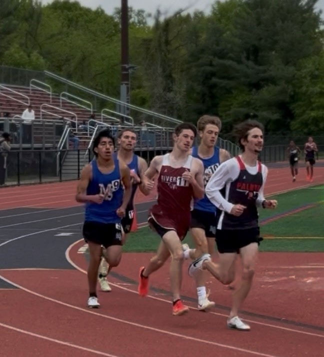 Athletes compete in a track race, running on a red track with spectators in the background. Palmyra is in the lead