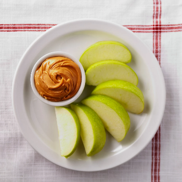 A white plate holds sliced green apples and a small bowl of creamy peanut butter.