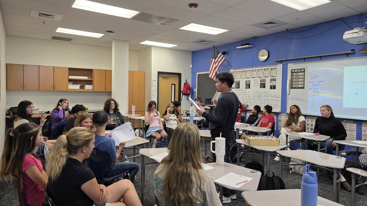 A young man stands and reads from papers to a classroom of seated students.
