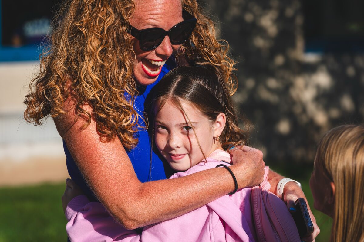 A smiling woman with curly hair and sunglasses hugs a young girl wearing a pink jacket.
