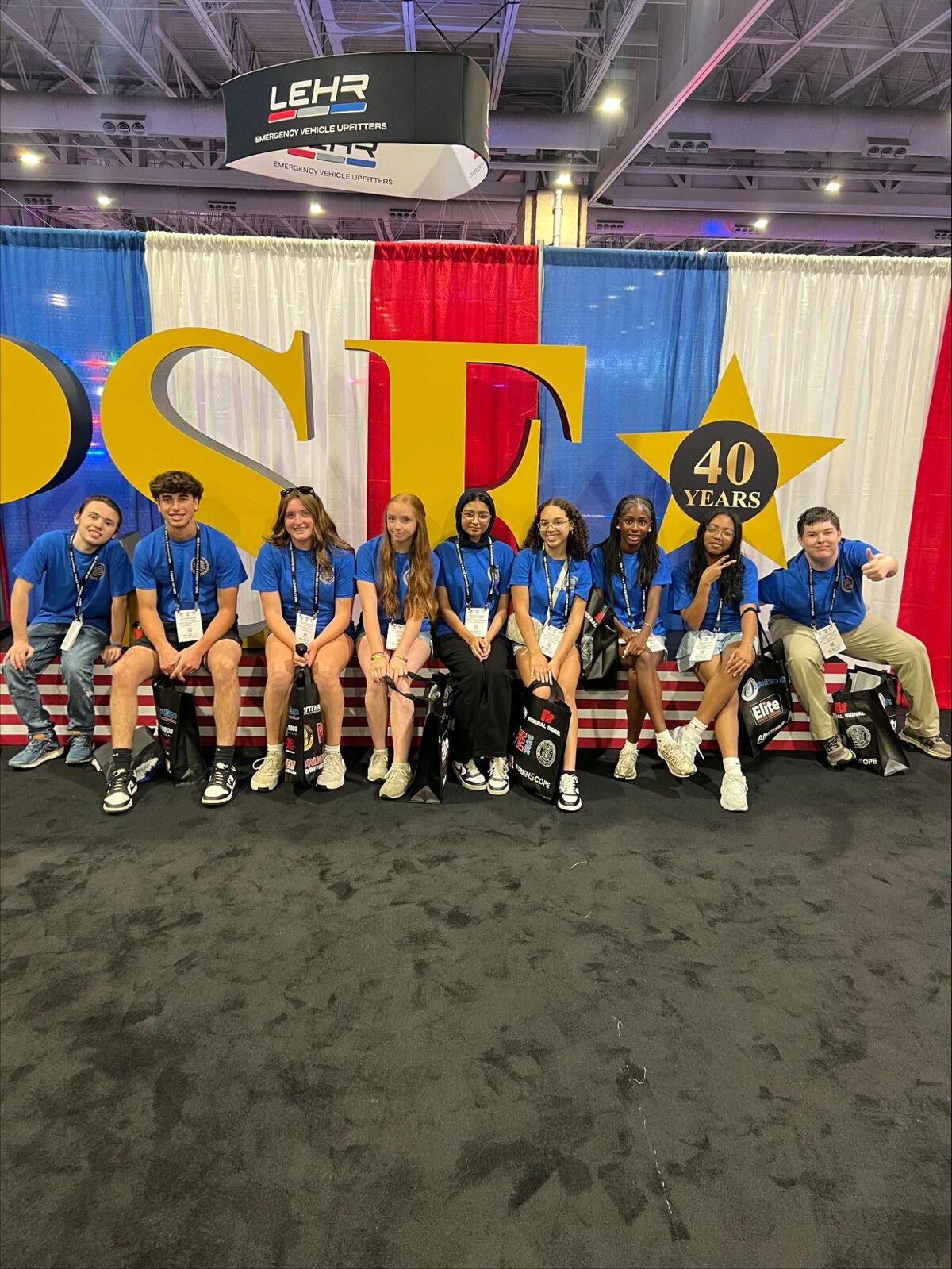 A group of young people in matching blue shirts sit on a bench in front of a large yellow 'PSE' sign and a red, white, and blue backdrop.
