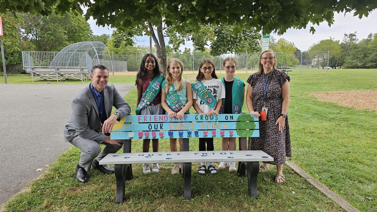 A group of four Girl Scouts and two adults proudly pose behind a colorful painted bench that reads "Friendship Grows on Our Buddy Bench".