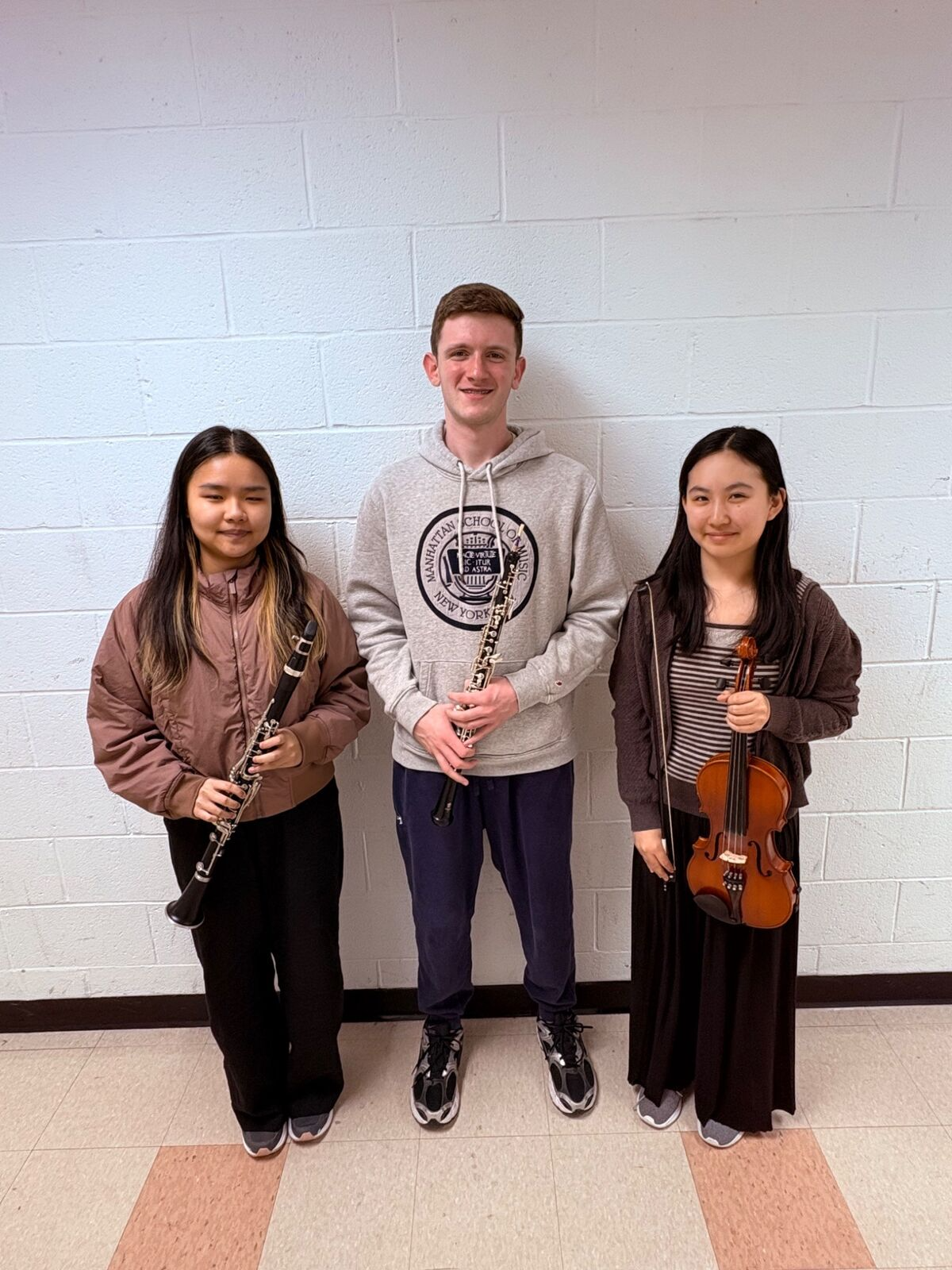 Three young musicians stand together, holding a clarinet, an oboe, and a violin.