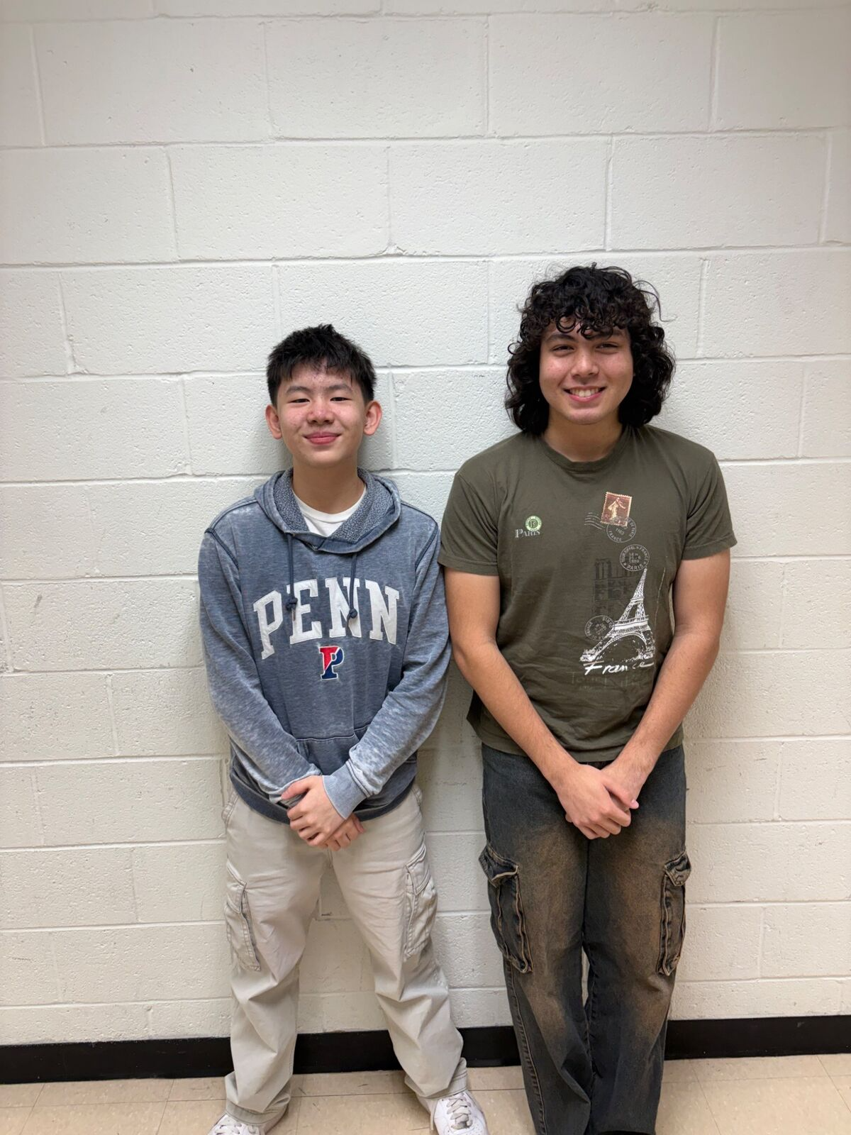 Two young men stand side-by-side against a white brick wall.