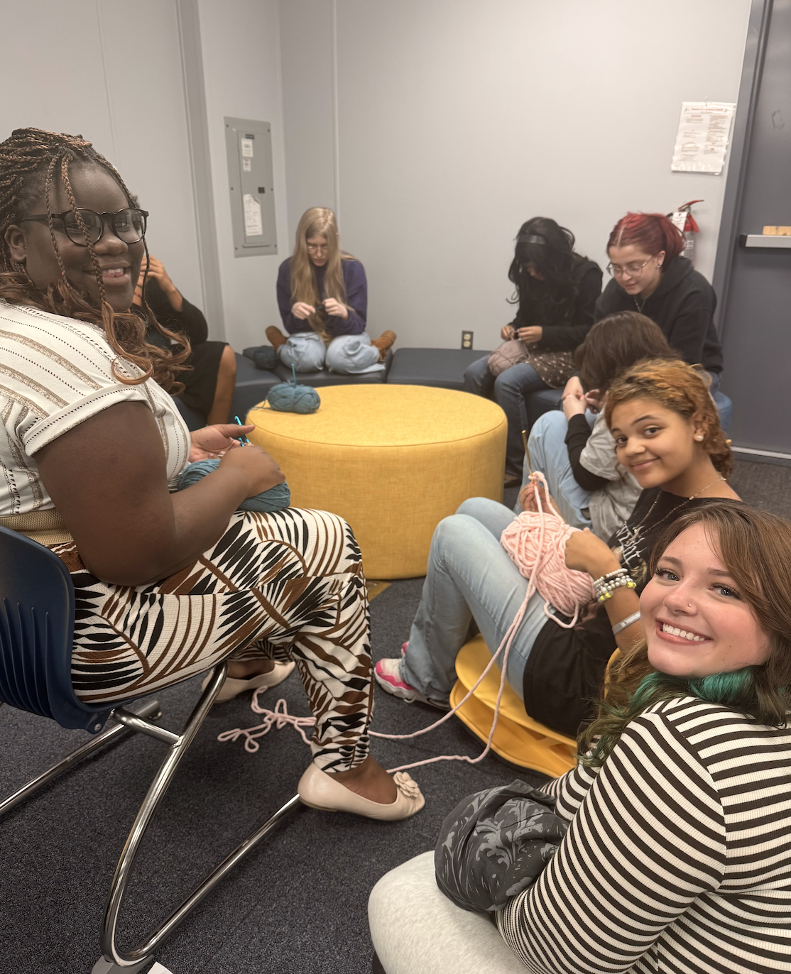 A group of people sit in a circle in a room, knitting or crocheting together.