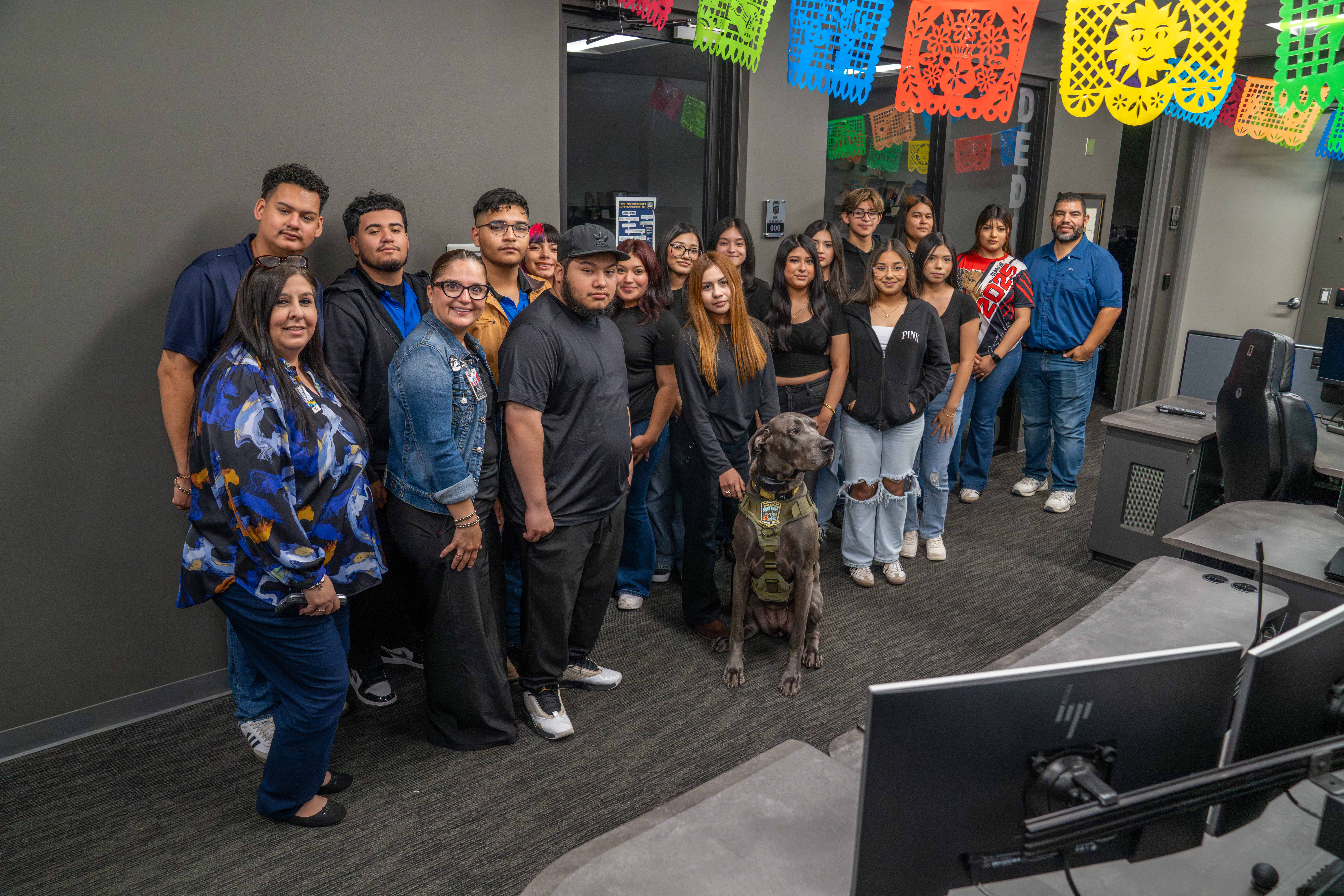A diverse group of people and a dog stand together in an office decorated with colorful papel picado banners.