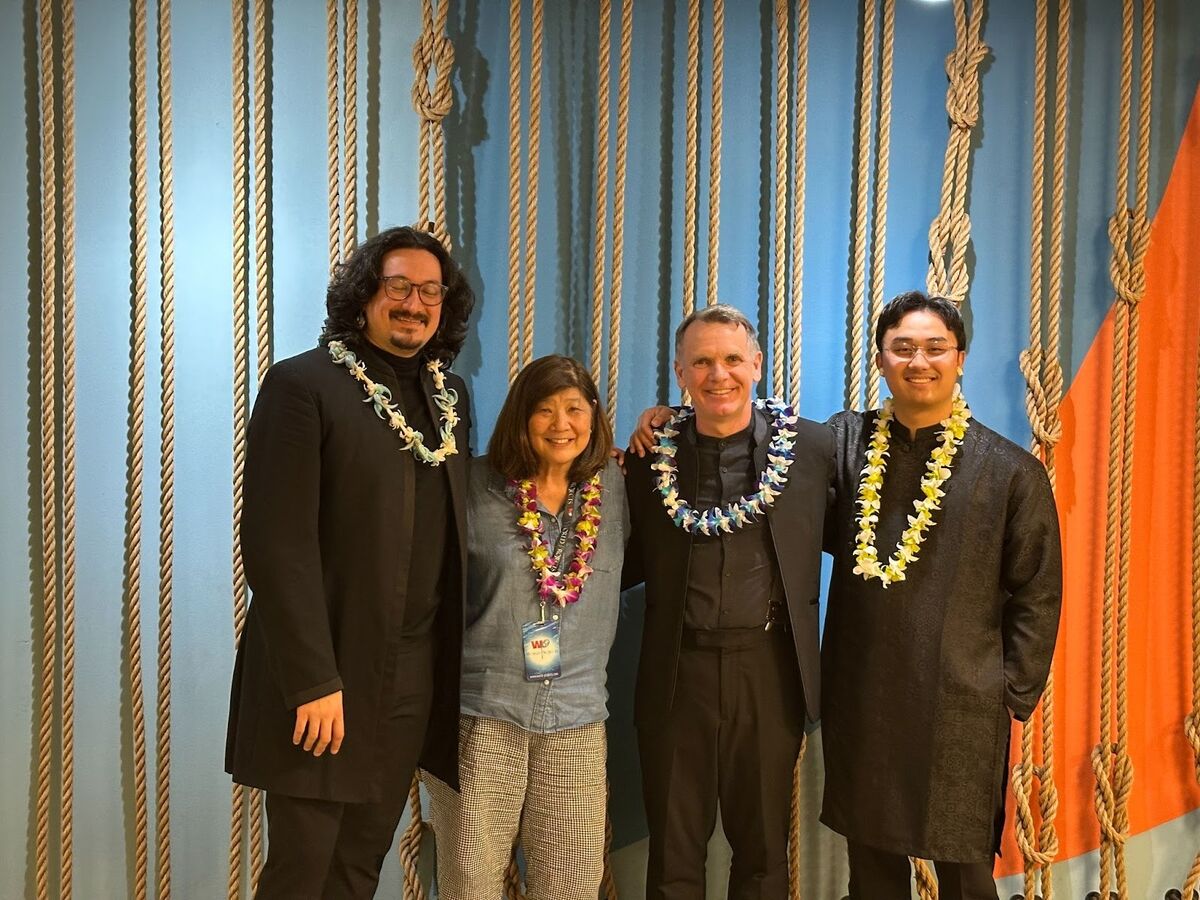 Four people stand together, wearing colorful leis and smiling in front of a backdrop of ropes and blue fabric.