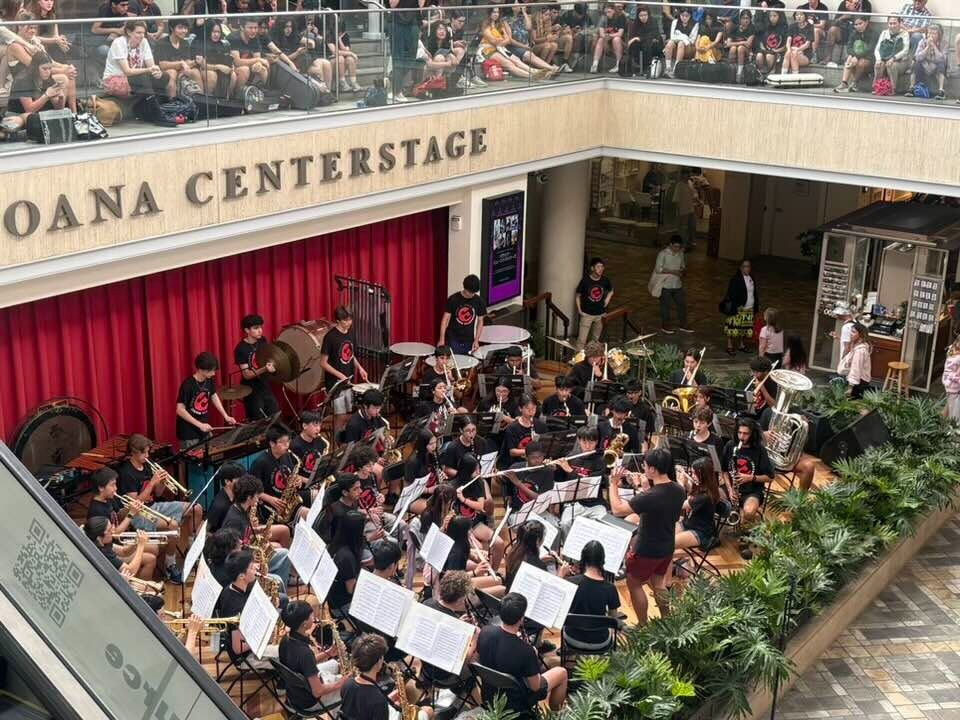 A large ensemble of young musicians plays instruments on a stage in a shopping center.