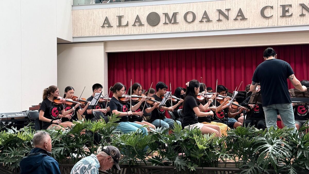 A youth orchestra performs with a conductor in front of a red curtain and the text "ALA MOANA CENTER".
