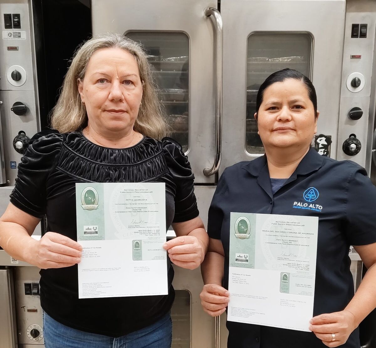 Two women stand in front of commercial ovens, each holding a Food Safety Manager certification.