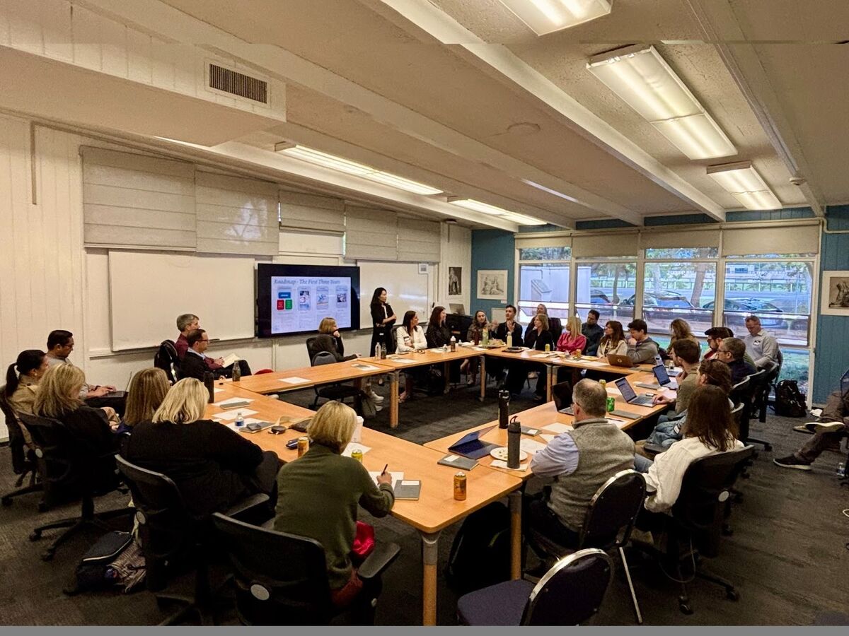A diverse group of people are seated around a U-shaped table in a conference room, listening to a presenter.