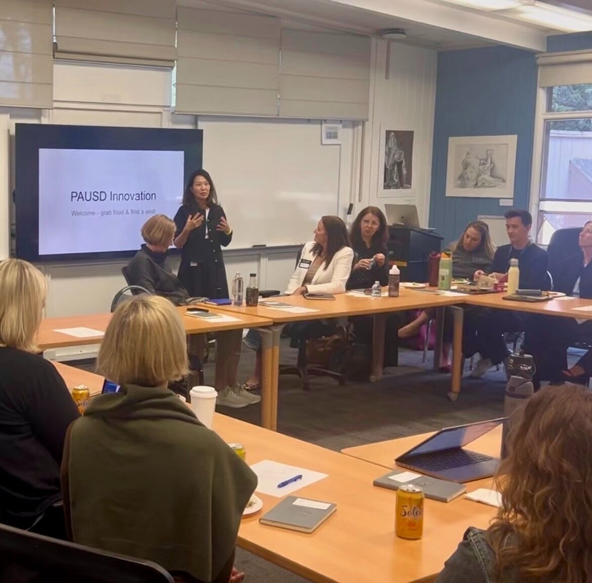 A woman presents to a group of people seated around a U-shaped table in a conference room.