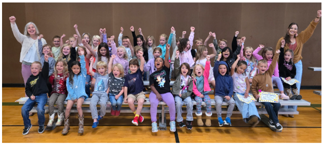 A large group of excited children and two adults pose with raised fists in a gymnasium.