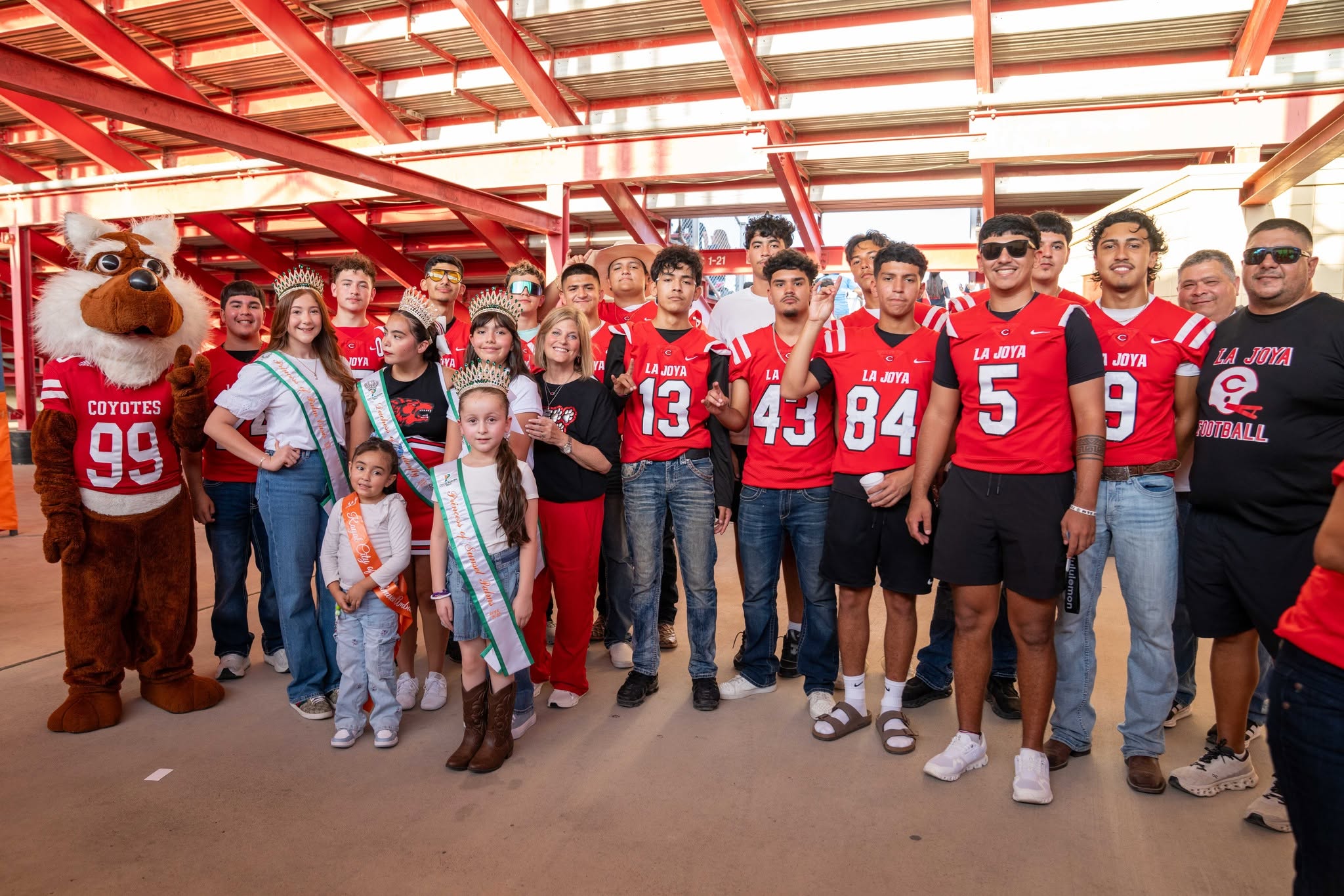 A group of people, including a mascot, football players, and young girls wearing sashes and crowns, pose for a photo under stadium bleachers.