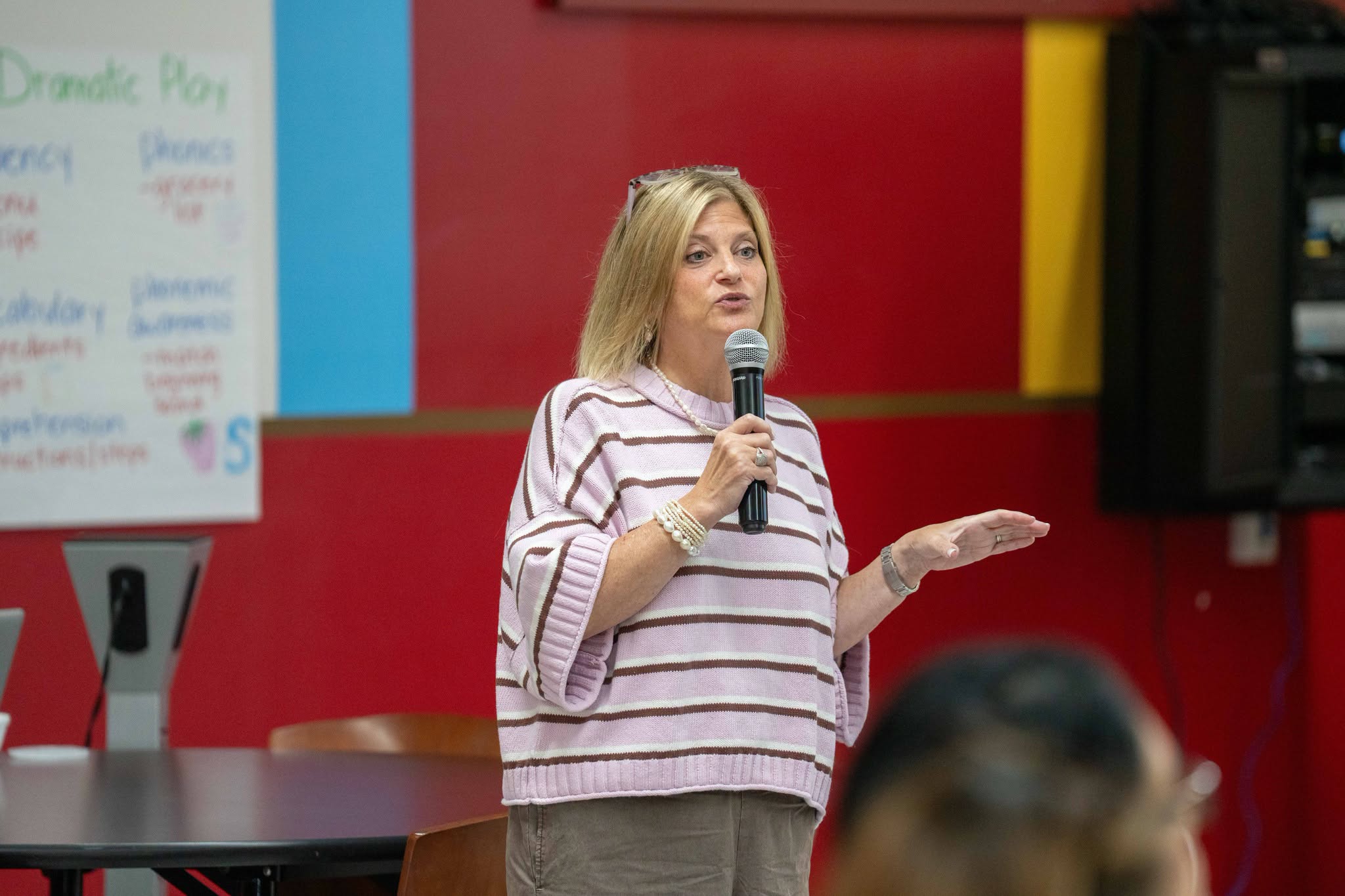 A woman with blonde hair speaks into a microphone, gesturing with her hands.