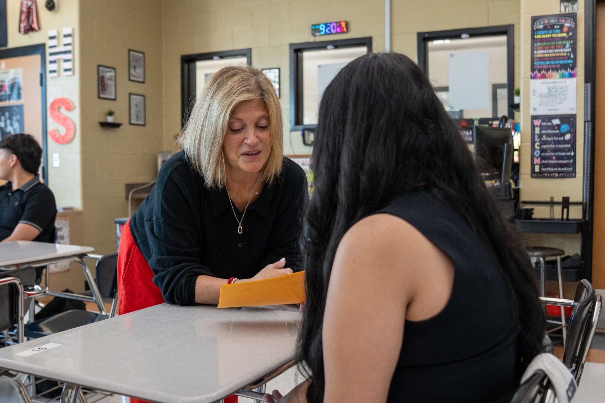 A teacher leans over a desk, speaking to a student in a classroom.
