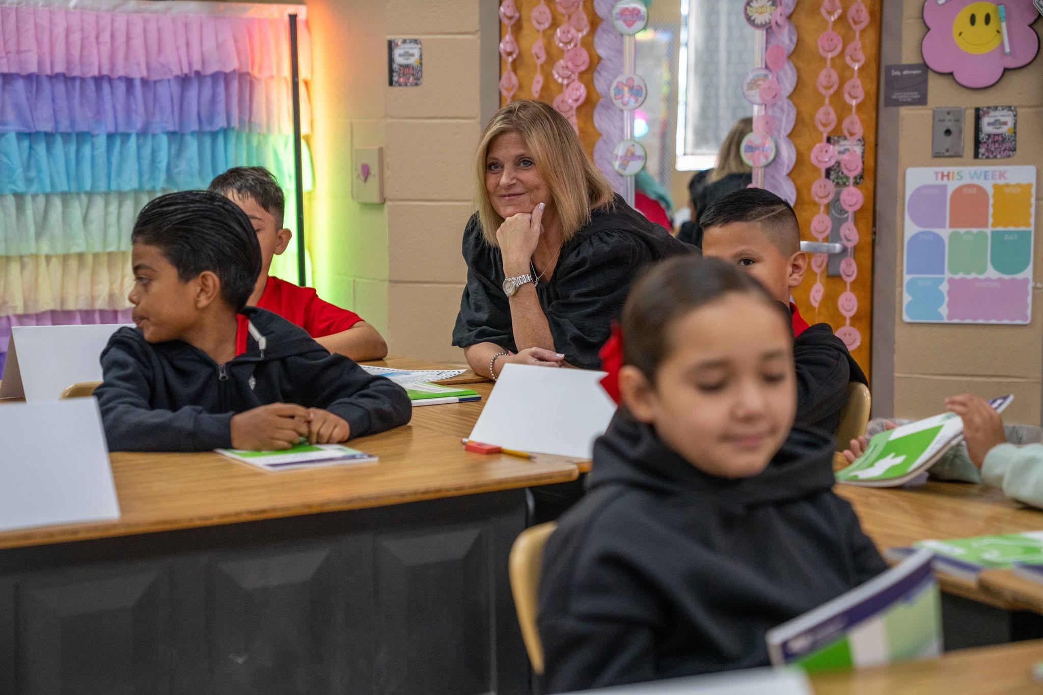 A teacher smiles while observing students at their desks in a colorful classroom.