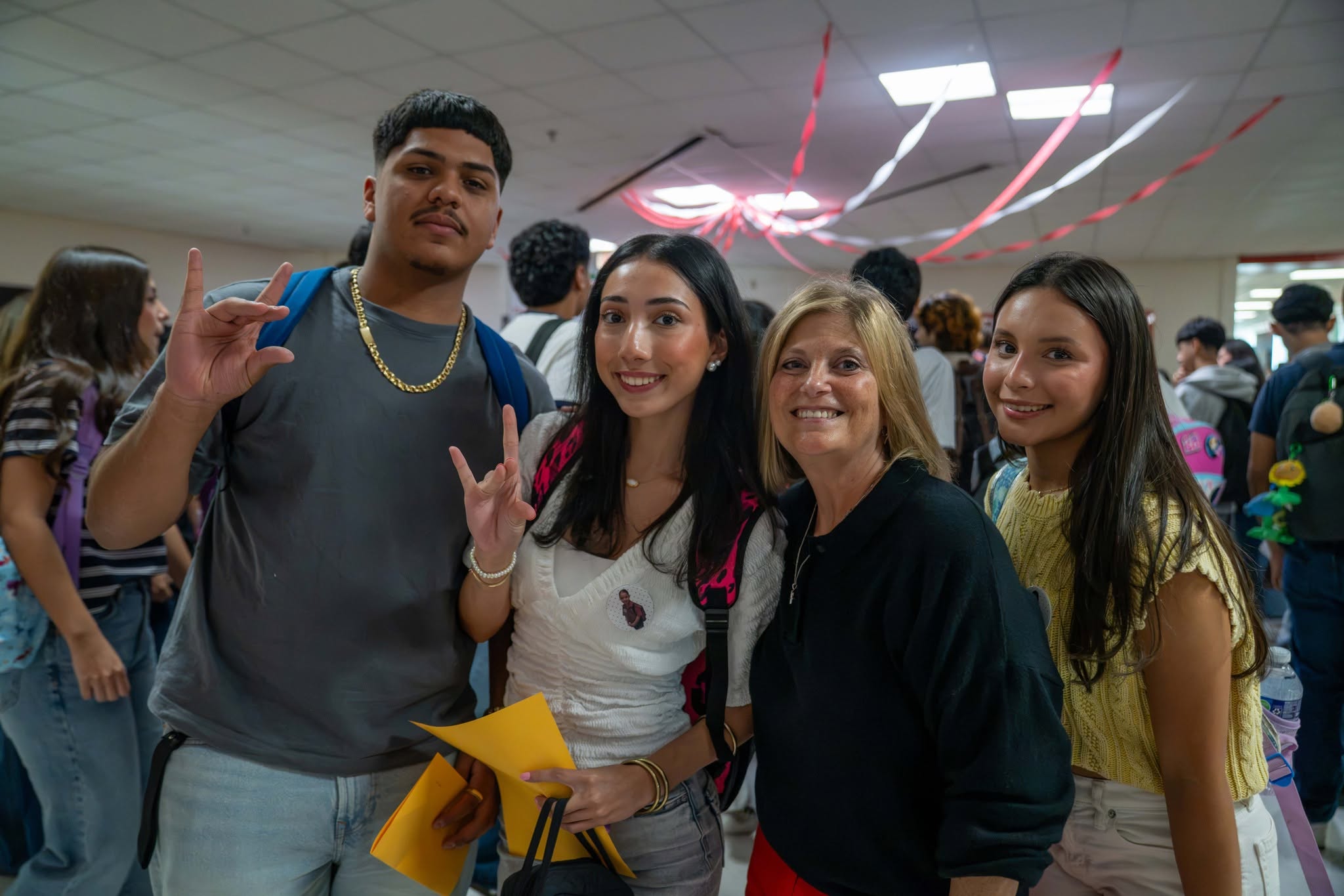 A group of smiling students and an adult pose for a photo in a hallway decorated with red and white streamers.
